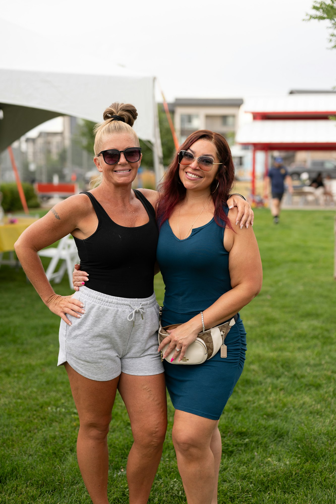 Two people smiling outdoors with sunglasses, standing on grass, with tents and buildings in the background.