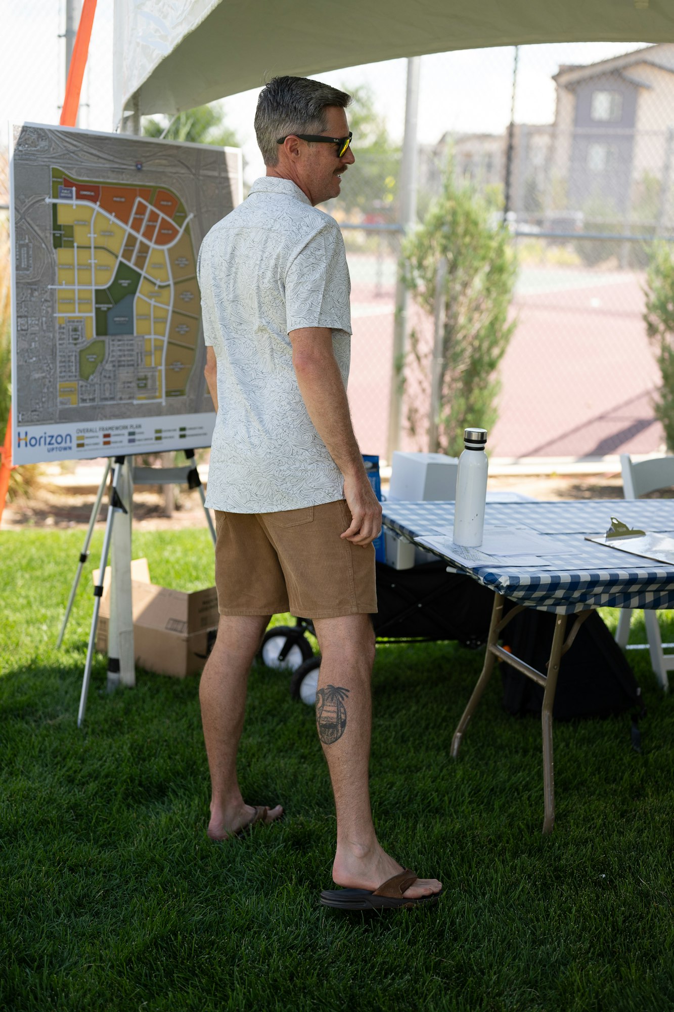 A man in casual attire stands near a map under a canopy, with a table and a water bottle nearby.