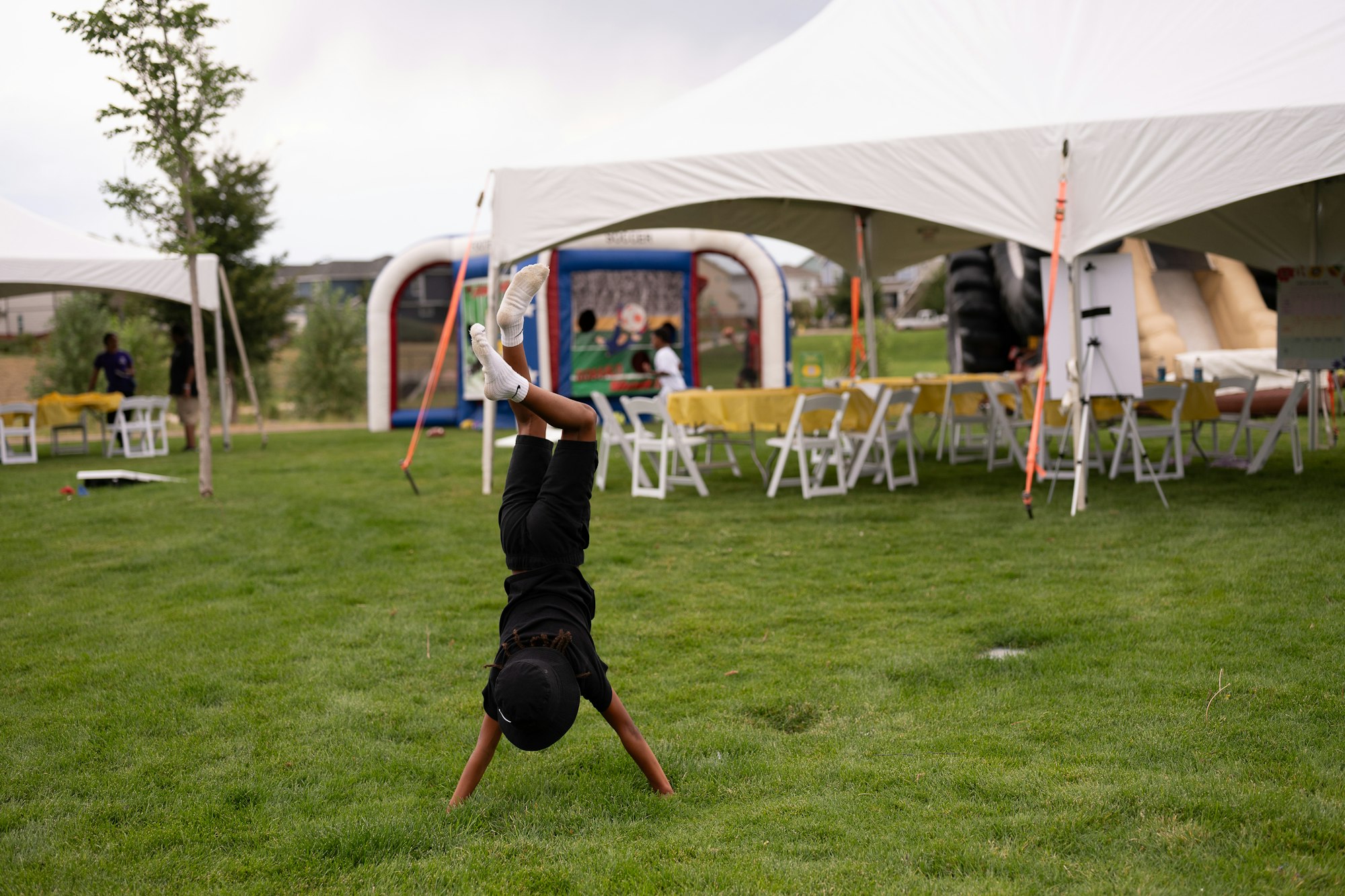 A child doing a handstand on grass near outdoor tents with tables and chairs set up for an event.