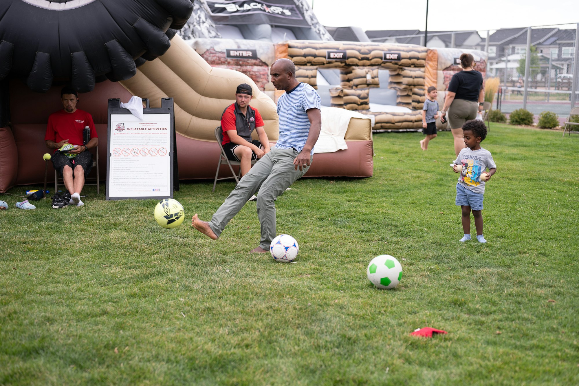 A man playing soccer barefoot on grass with kids and a bouncy house in the background.