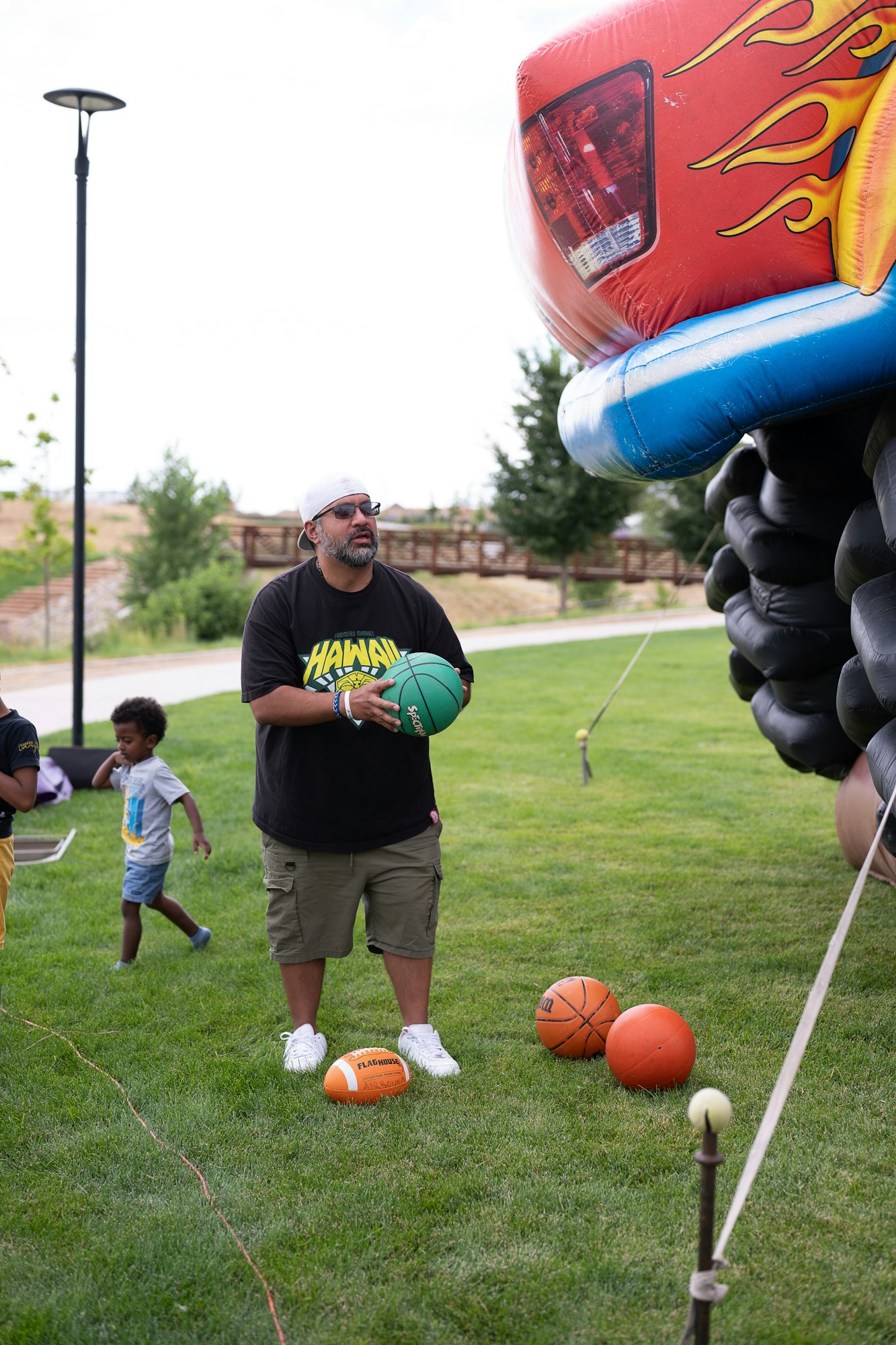 A man holds a green basketball near an inflatable car with flames, while children play on the grass nearby.