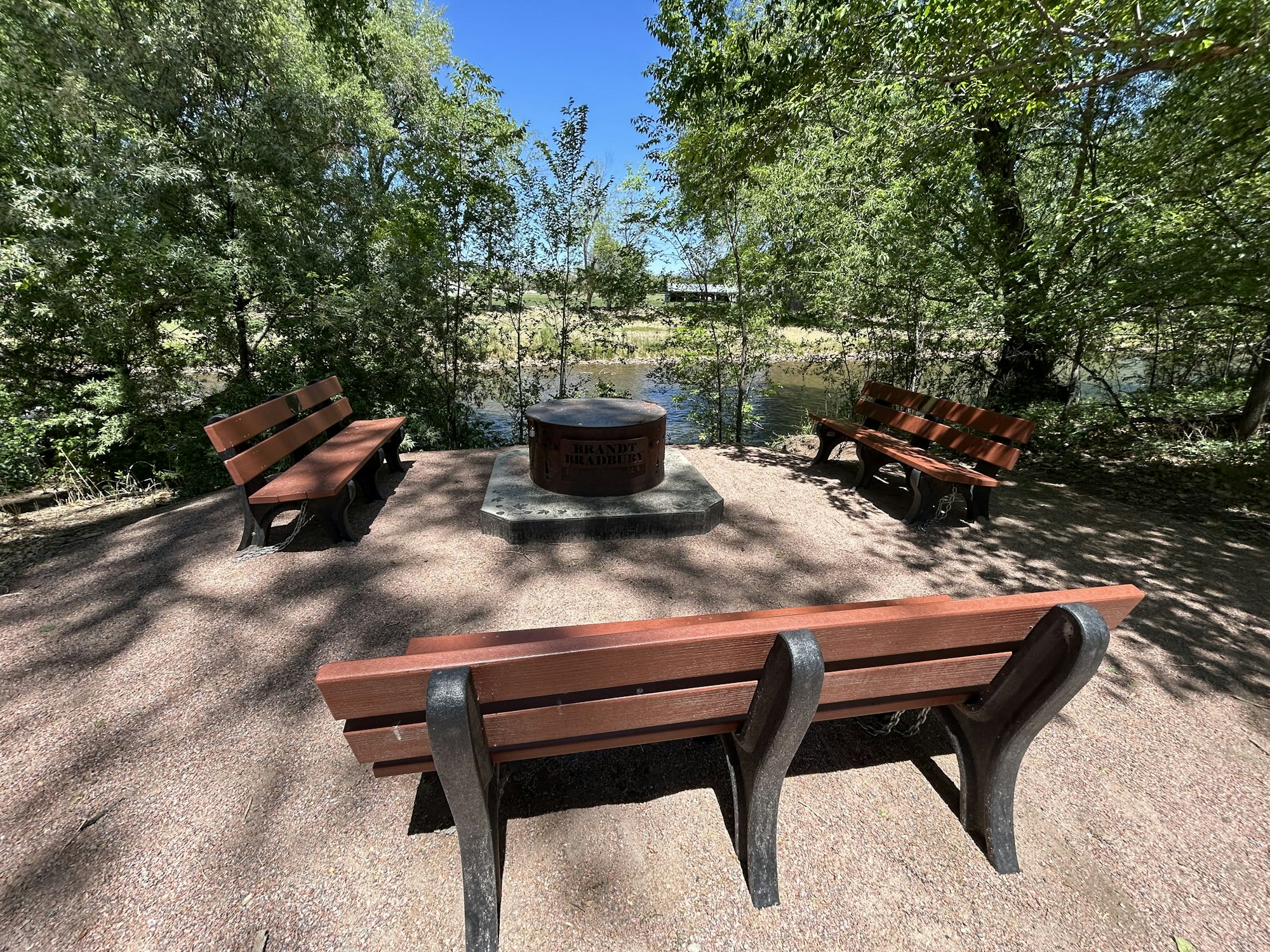 A peaceful spot with benches arranged around a fire ring, surrounded by trees and near a body of water. Ideal for gatherings.
