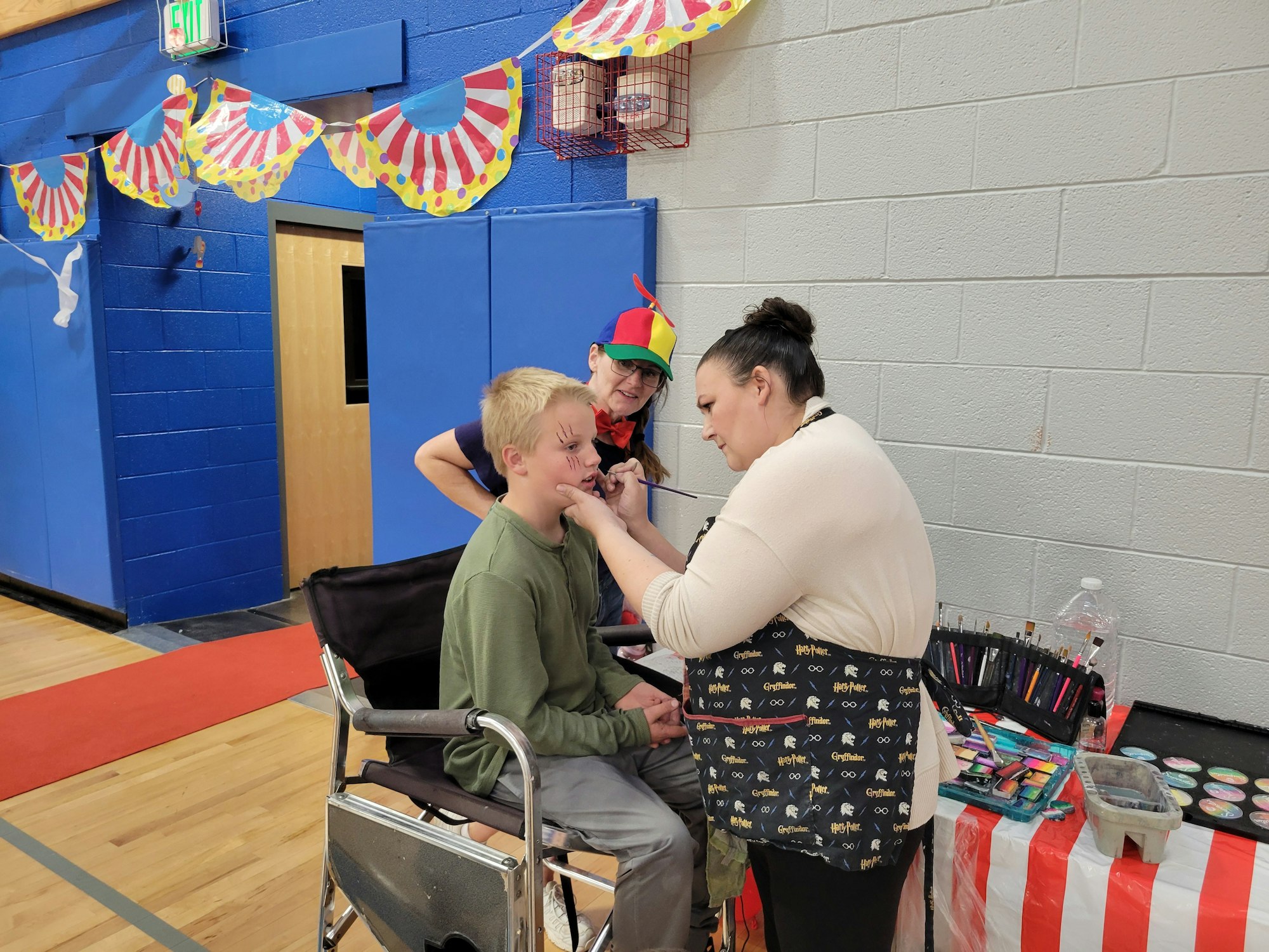 A boy is getting his face painted at a festive event, while two women assist him, surrounded by colorful decorations.