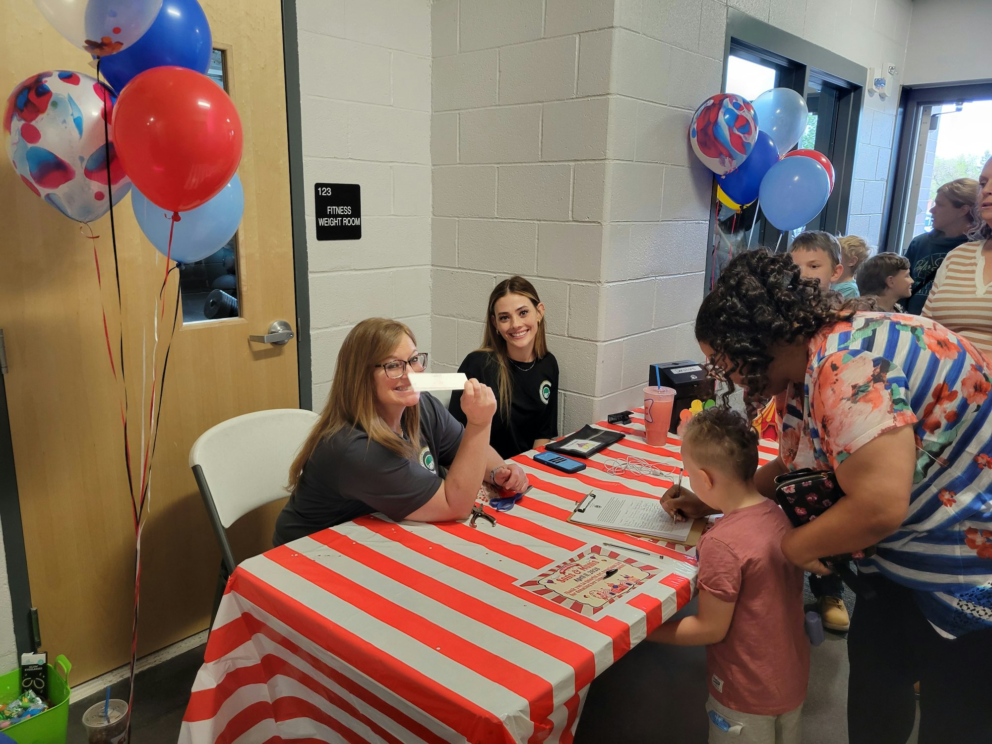 Two women are at a table with balloons, greeting visitors, while a child and an adult fill out a form at a festive event.