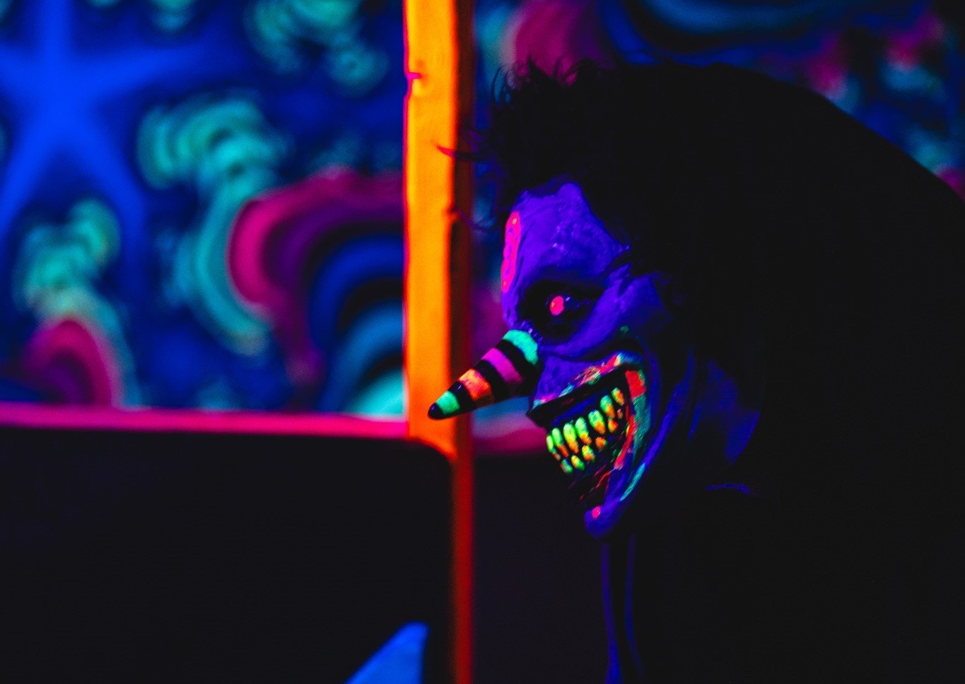 A glowing, creepy mask with a pointed nose and jagged grin in colorful neon light, set against a psychedelic background.