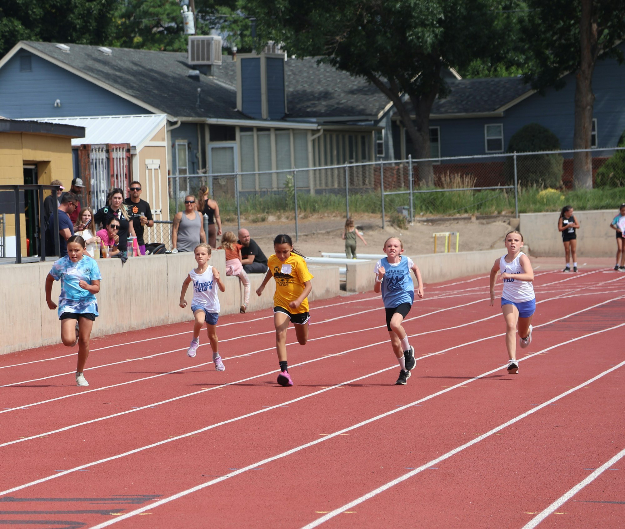 Kids are sprinting on a red track during a race, with spectators watching from the sidelines.
