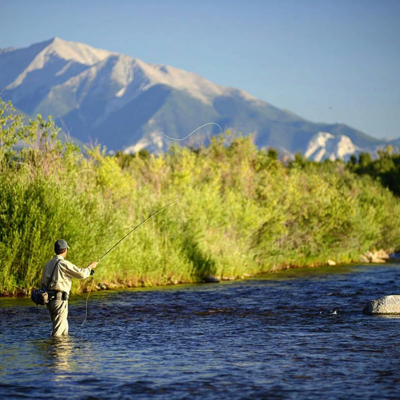 Person fly fishing in a river with mountains in the background.