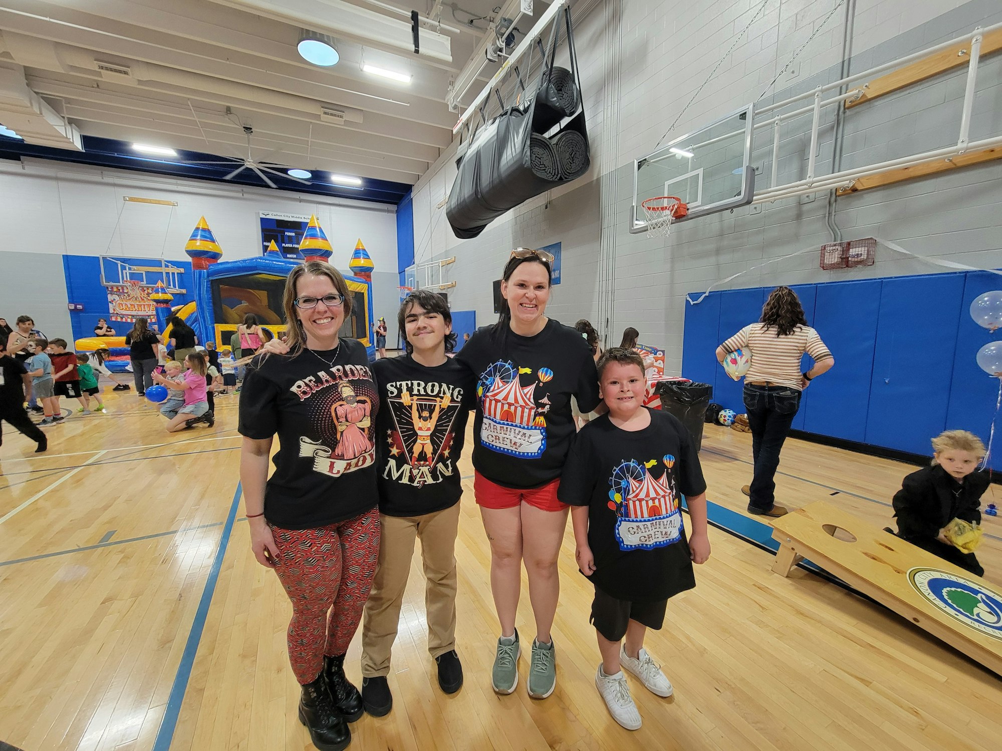 A group of four people wearing themed t-shirts poses together in a gym with kids playing and a bounce house in the background.