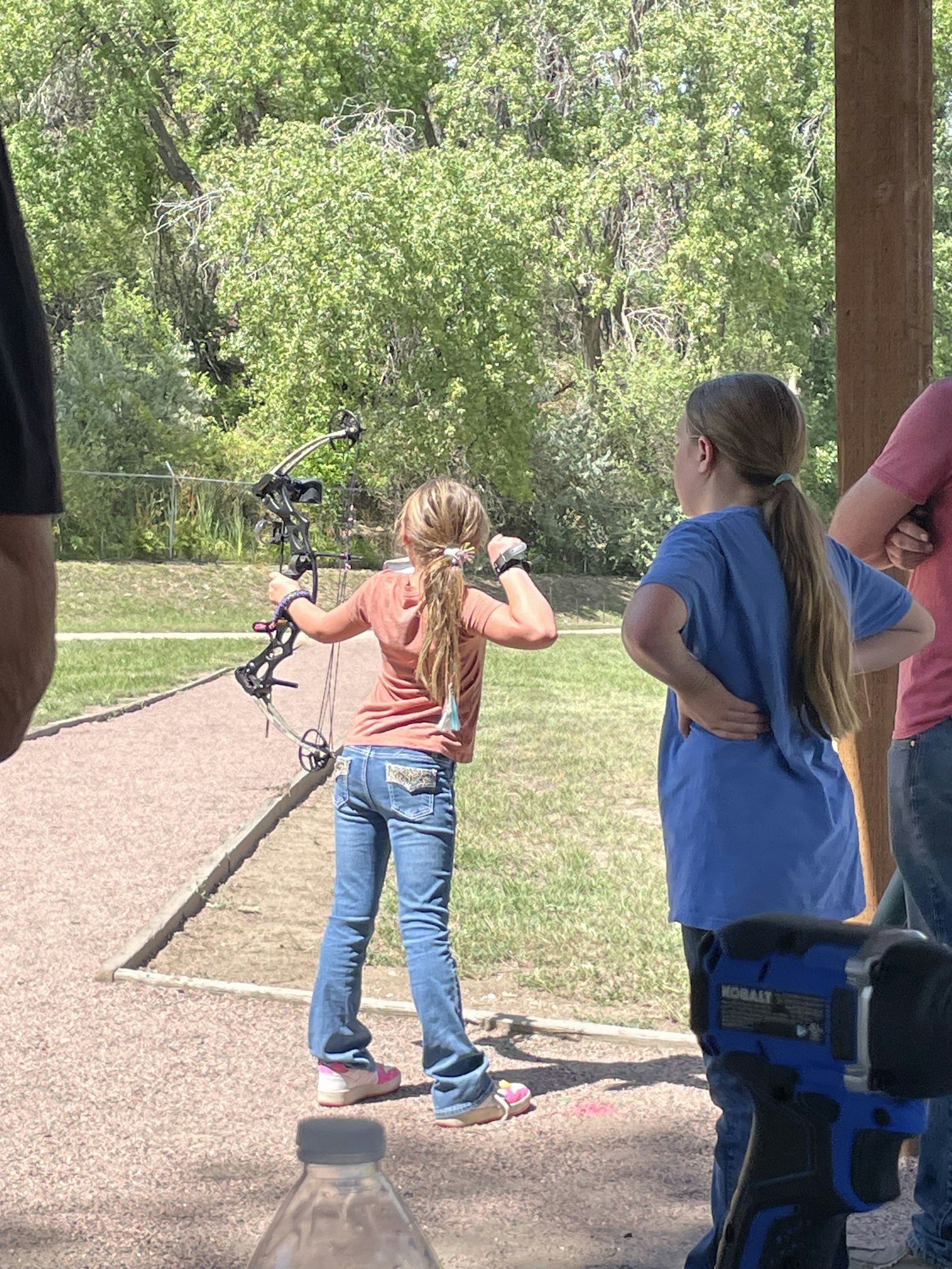 Two people practicing archery outdoors, with one person aiming a bow and another watching.