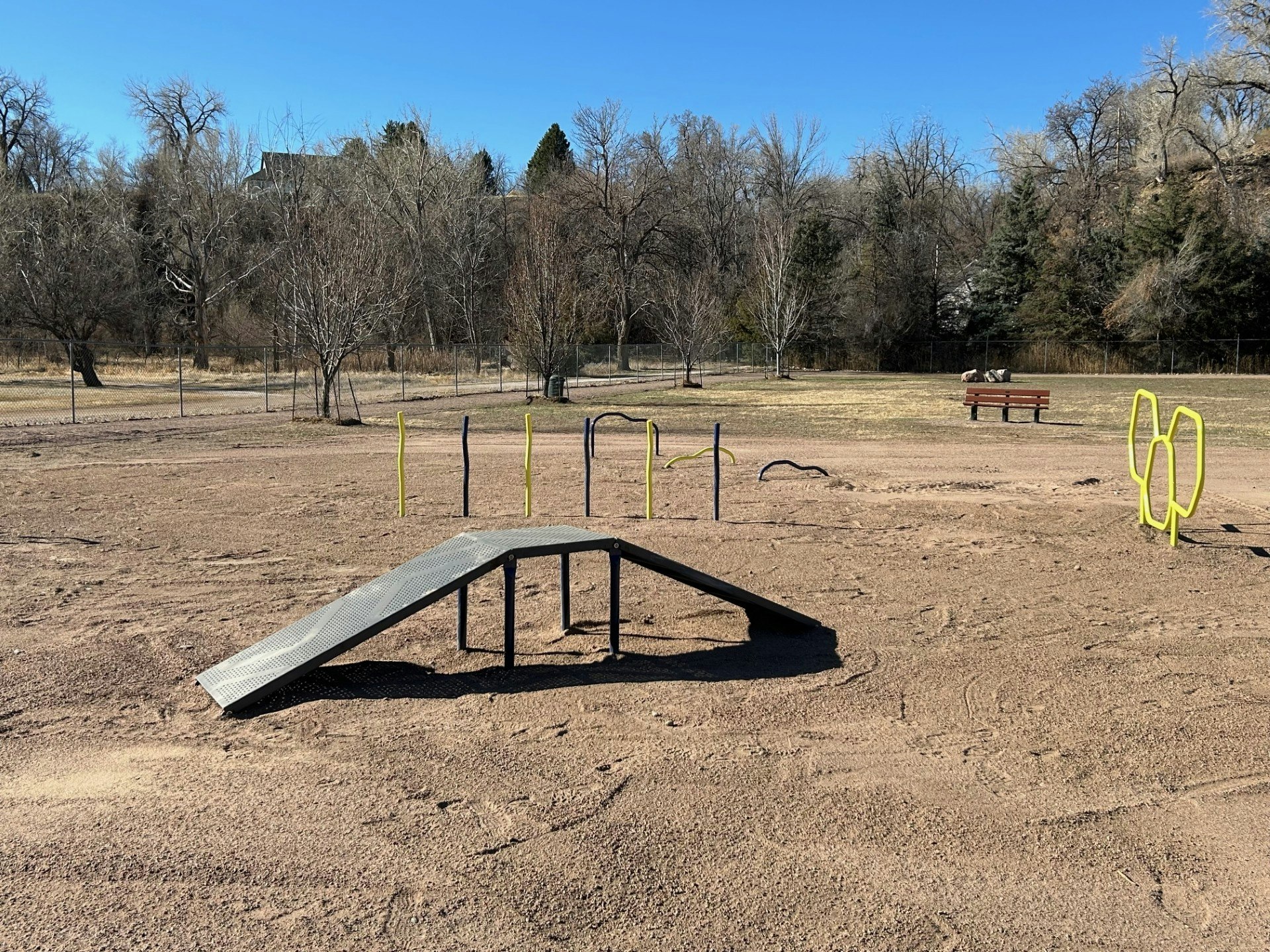 A dog park with agility equipment: ramp, hoops, and poles. Trees in the background, a bench with a person, under clear blue sky.