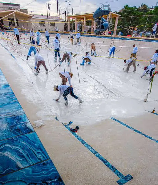 Workers are coating the bottom of an empty swimming pool, using tools and spray equipment in a collaborative effort.