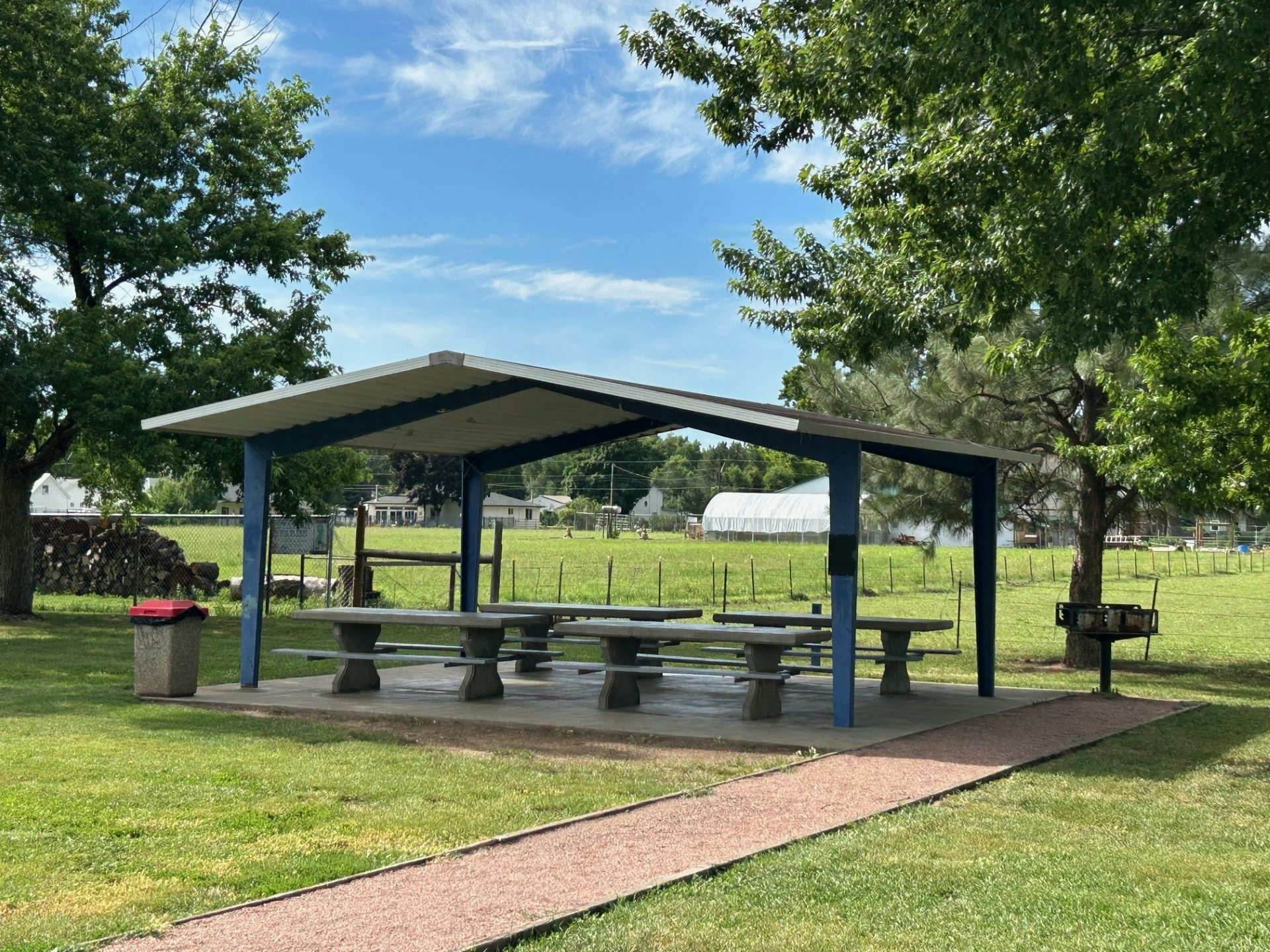 A picnic area with a shelter, tables, a trash can, a barbecue grill, and surrounding trees under a clear sky at Rouse Park