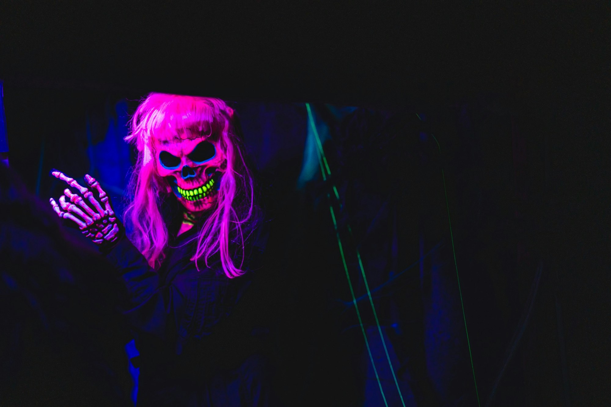 A figure in a neon skull mask and skeleton gloves under blacklight, with bright pink hair.