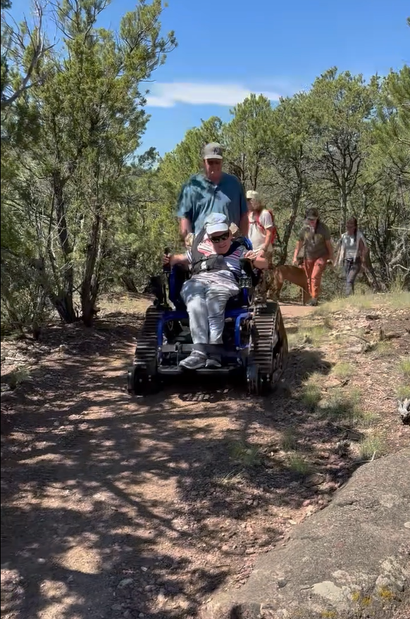 A person in a tracked wheelchair is being assisted on a trail, with others walking nearby in a natural setting.