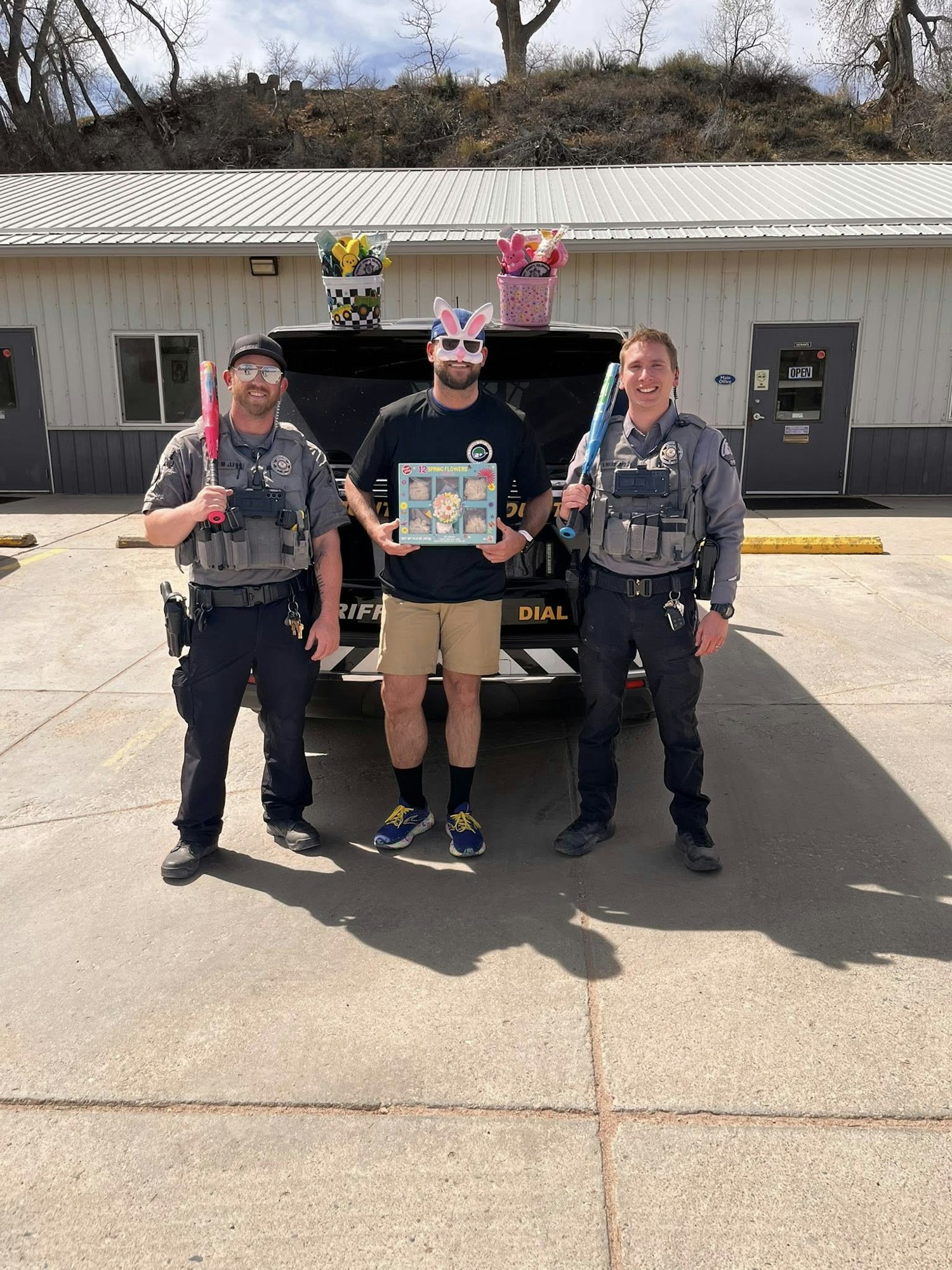 Three individuals, two in police uniforms and one with bunny ears, hold colorful items and smile in front of a vehicle.
