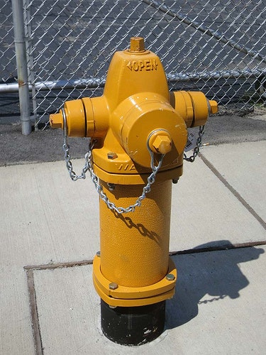 A yellow fire hydrant on a sidewalk near a chain-link fence.