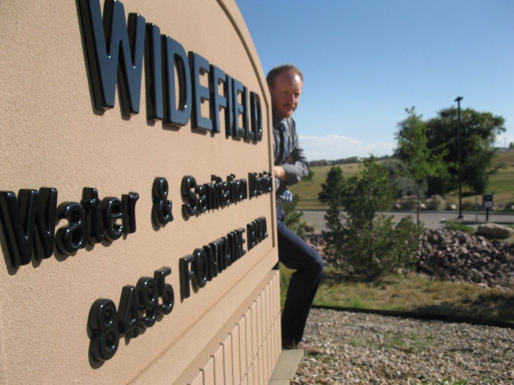 A person playfully poses behind a "Widefield Water & Sanitation" sign outdoors.