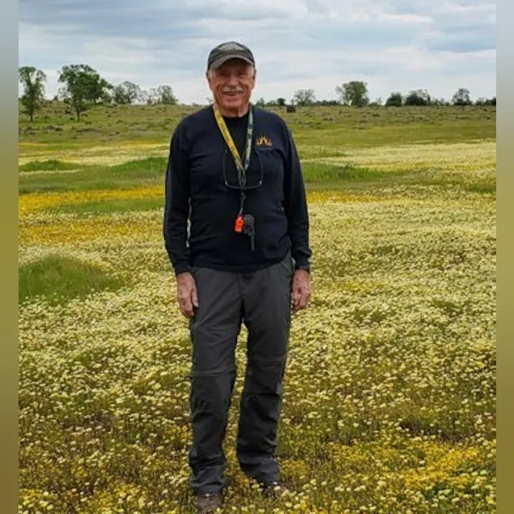 An older man stands in a field of yellow and white flowers, wearing casual outdoor clothing and a cap, with a scenic landscape behind him.