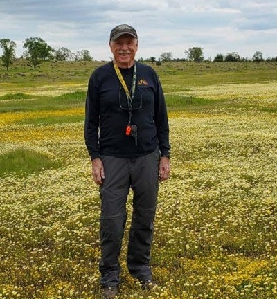 An older man stands in a field of yellow and white flowers, wearing casual outdoor clothing and a cap, with a scenic landscape behind him.