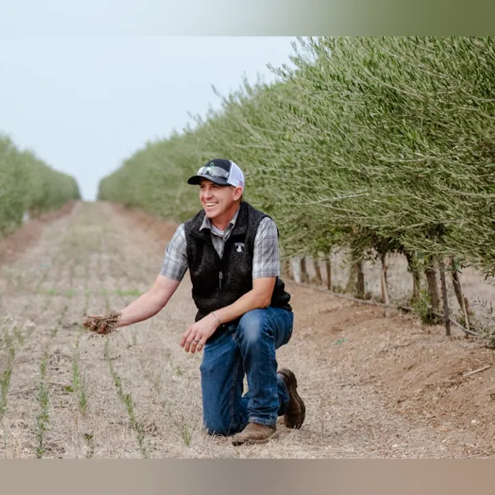 A smiling man kneels in a farm field, examining soil, surrounded by rows of green plants.