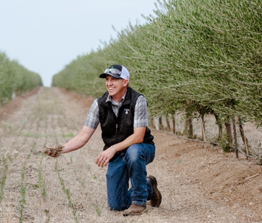 A smiling man kneels in a farm field, examining soil, surrounded by rows of green plants.