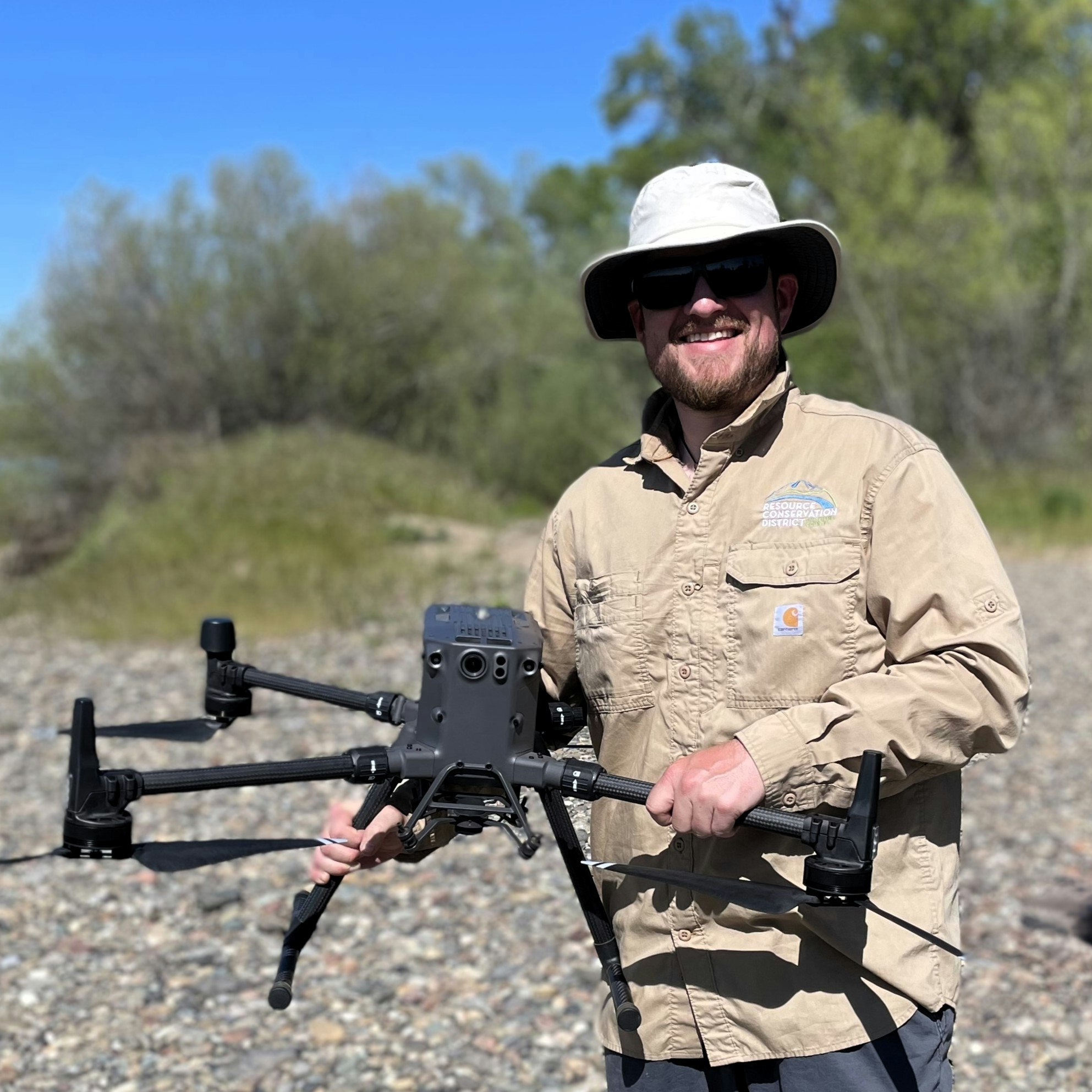 A smiling person holding a drone outdoors, wearing a hat and sunglasses.