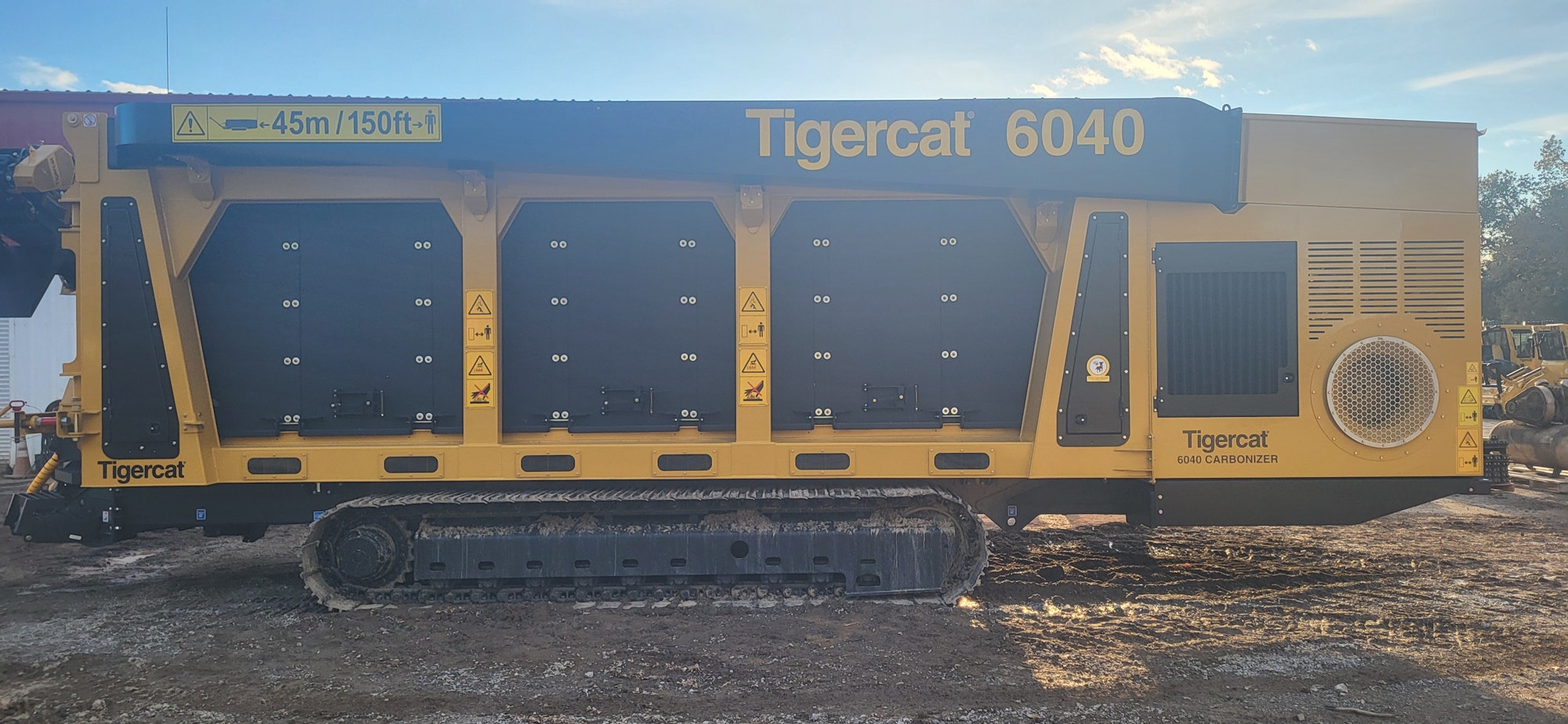 Yellow Tigercat 6040 carbonizer machine with black panels and warning signs on a dirt lot under blue sky.
