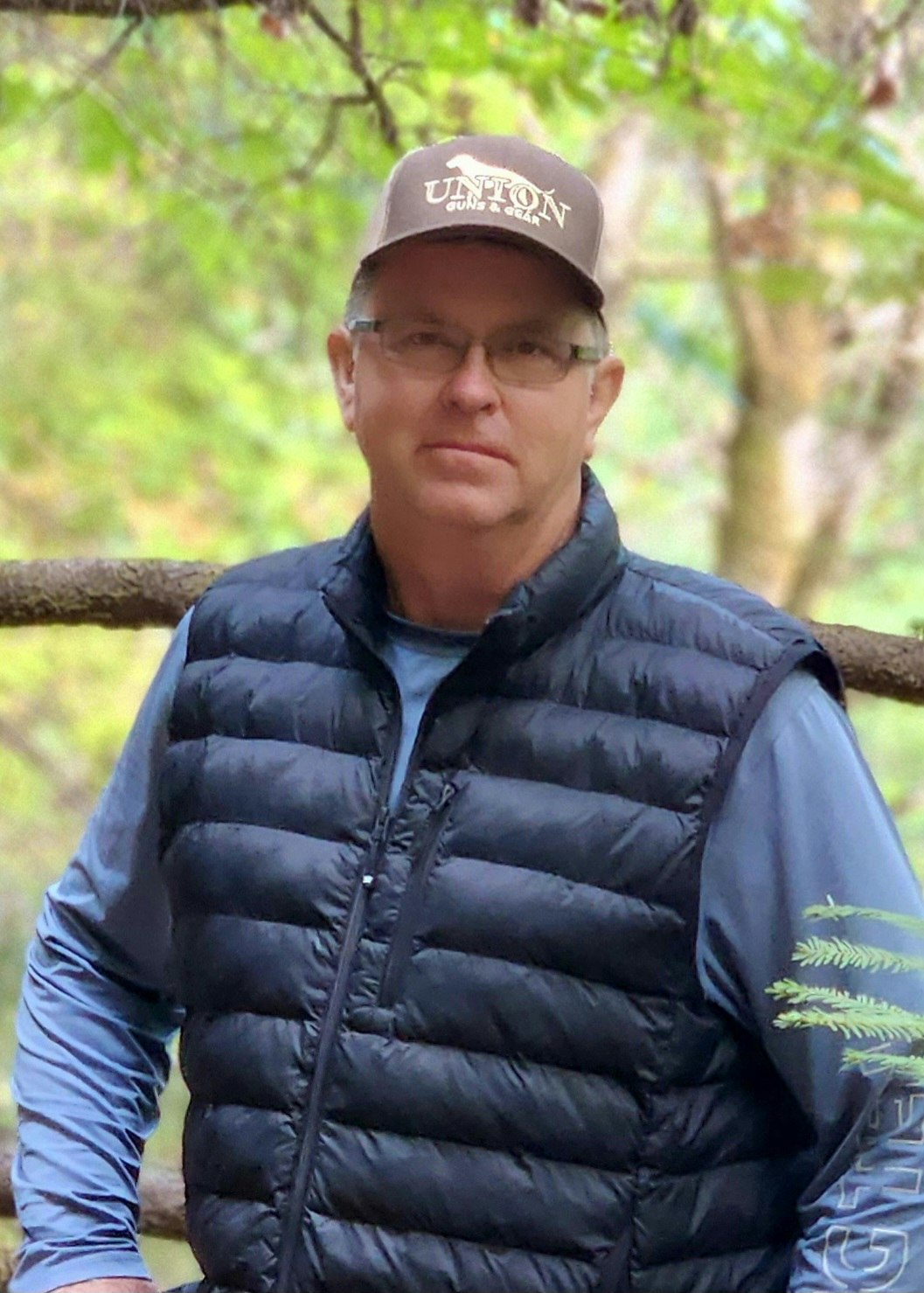 Man in a cap and black vest, standing outdoors with a blurred green background.