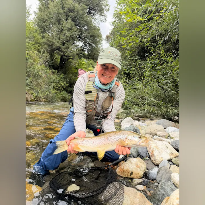 A person smiles while holding a large trout, standing in a stream surrounded by lush greenery and rocks.