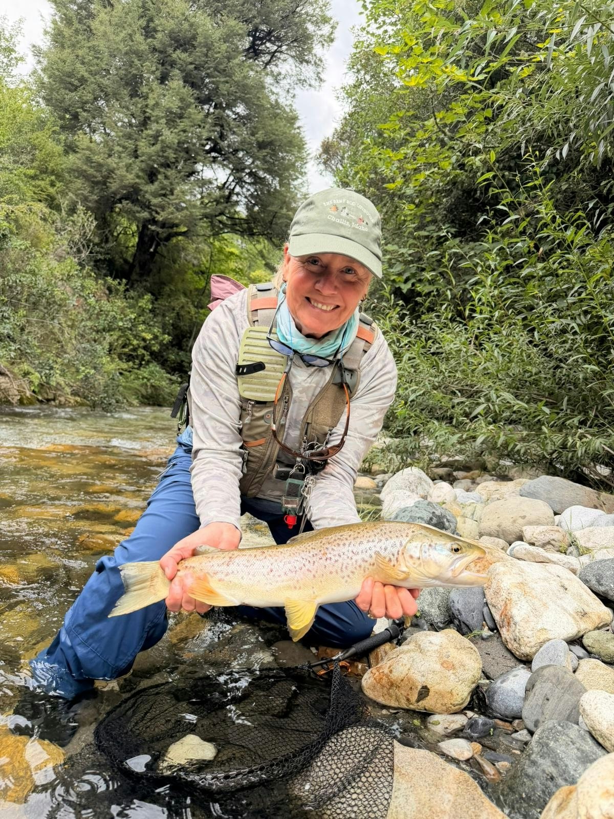 A person smiles while holding a large trout, standing in a stream surrounded by lush greenery and rocks.