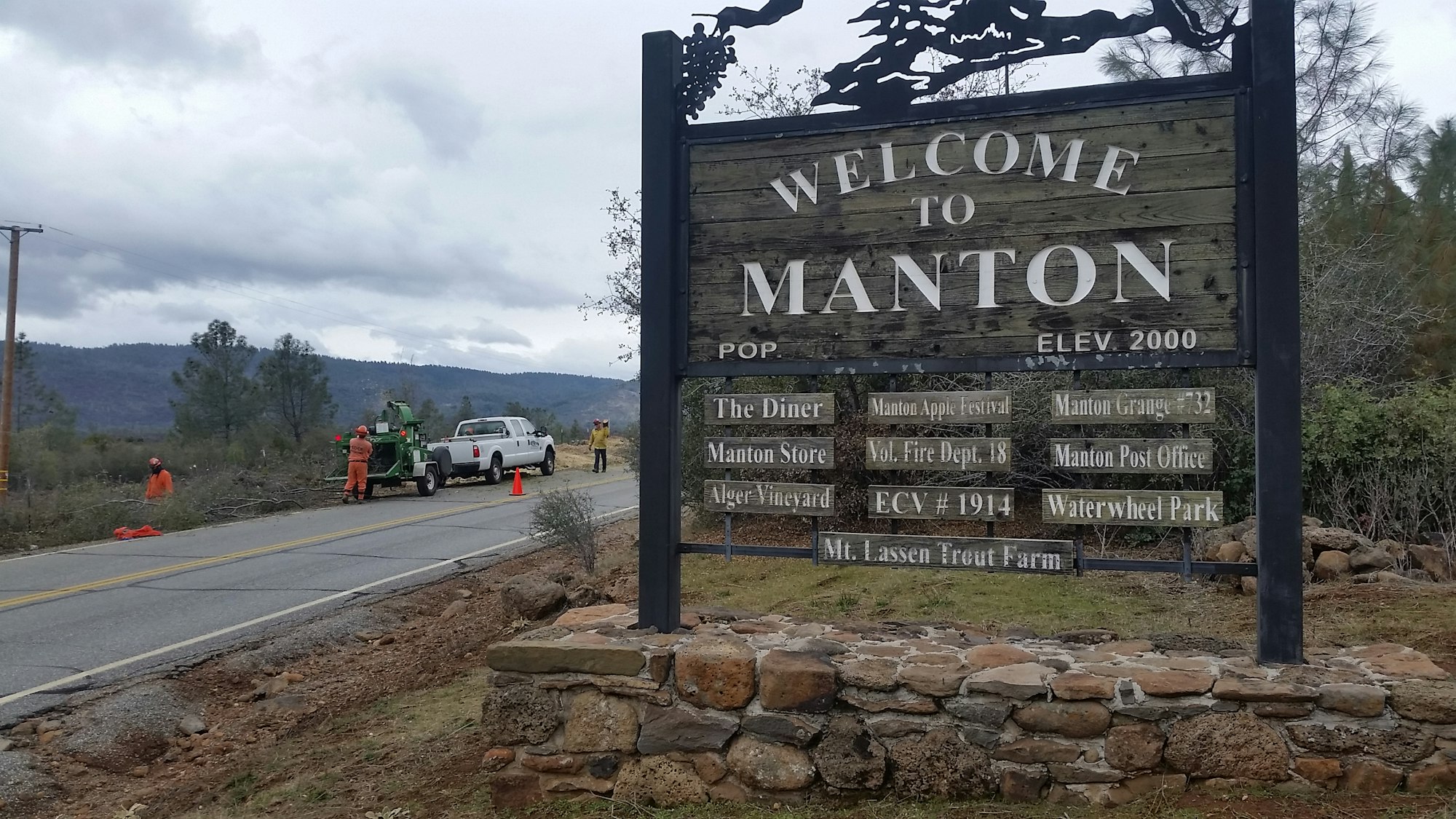 A welcome sign for Manton, CA, with local attractions listed, and workers performing maintenance near the road.