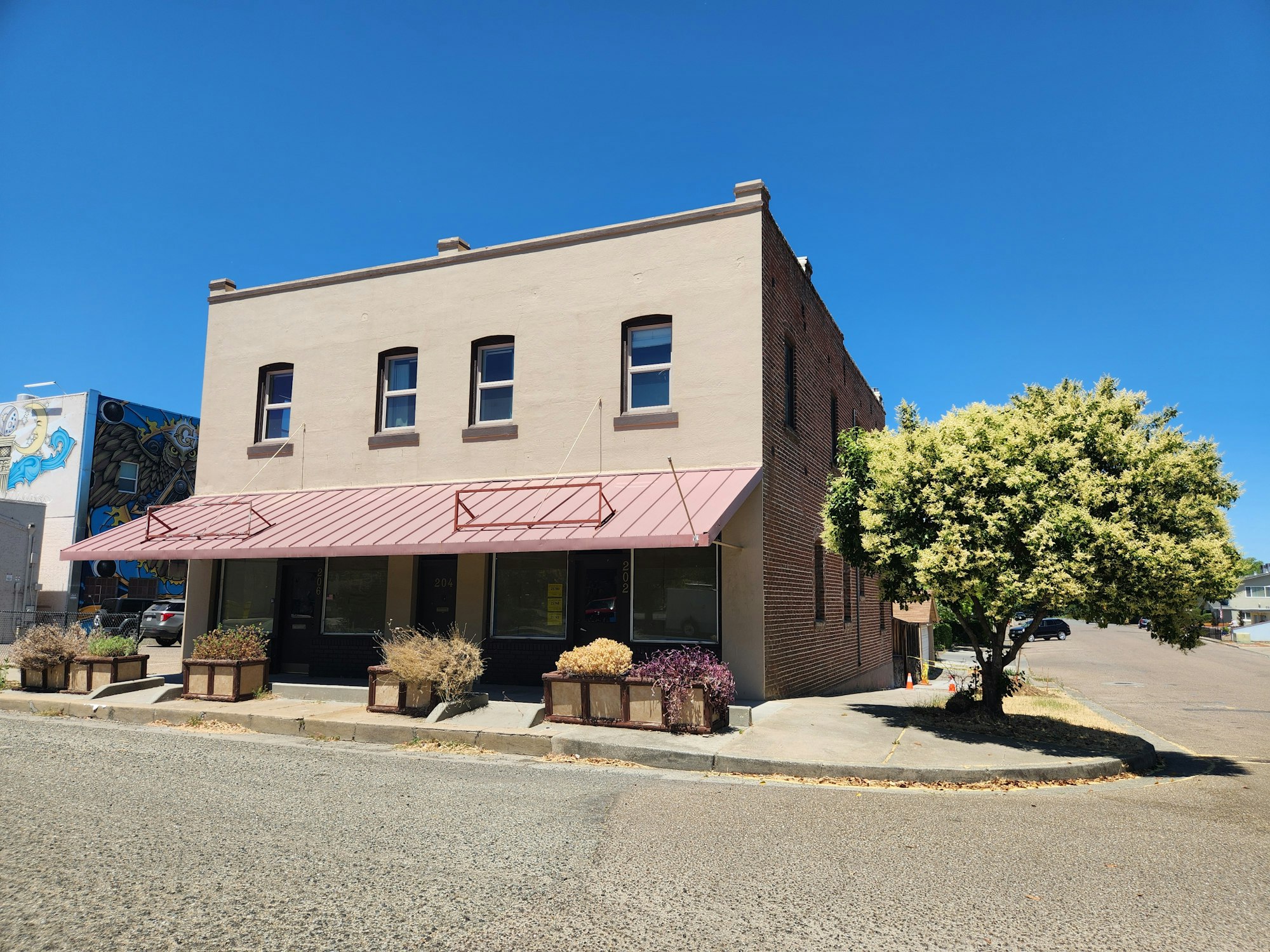 Corner building with awning, brick siding, a tree, blue sky, and a mural on adjacent building.