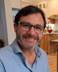 A smiling person with glasses in a casual blue shirt, indoors.