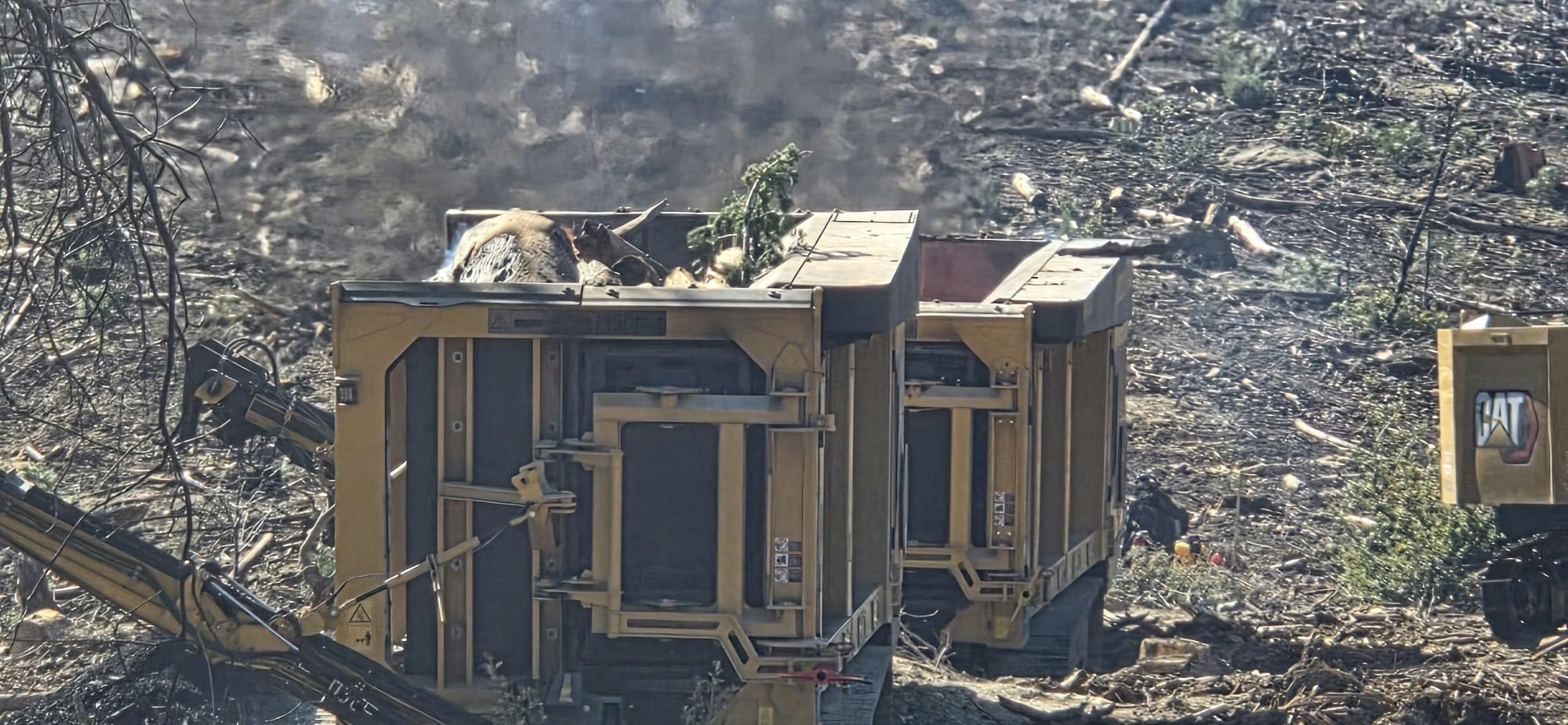 The image shows heavy machinery working in a deforested area, with smoke and debris visible.