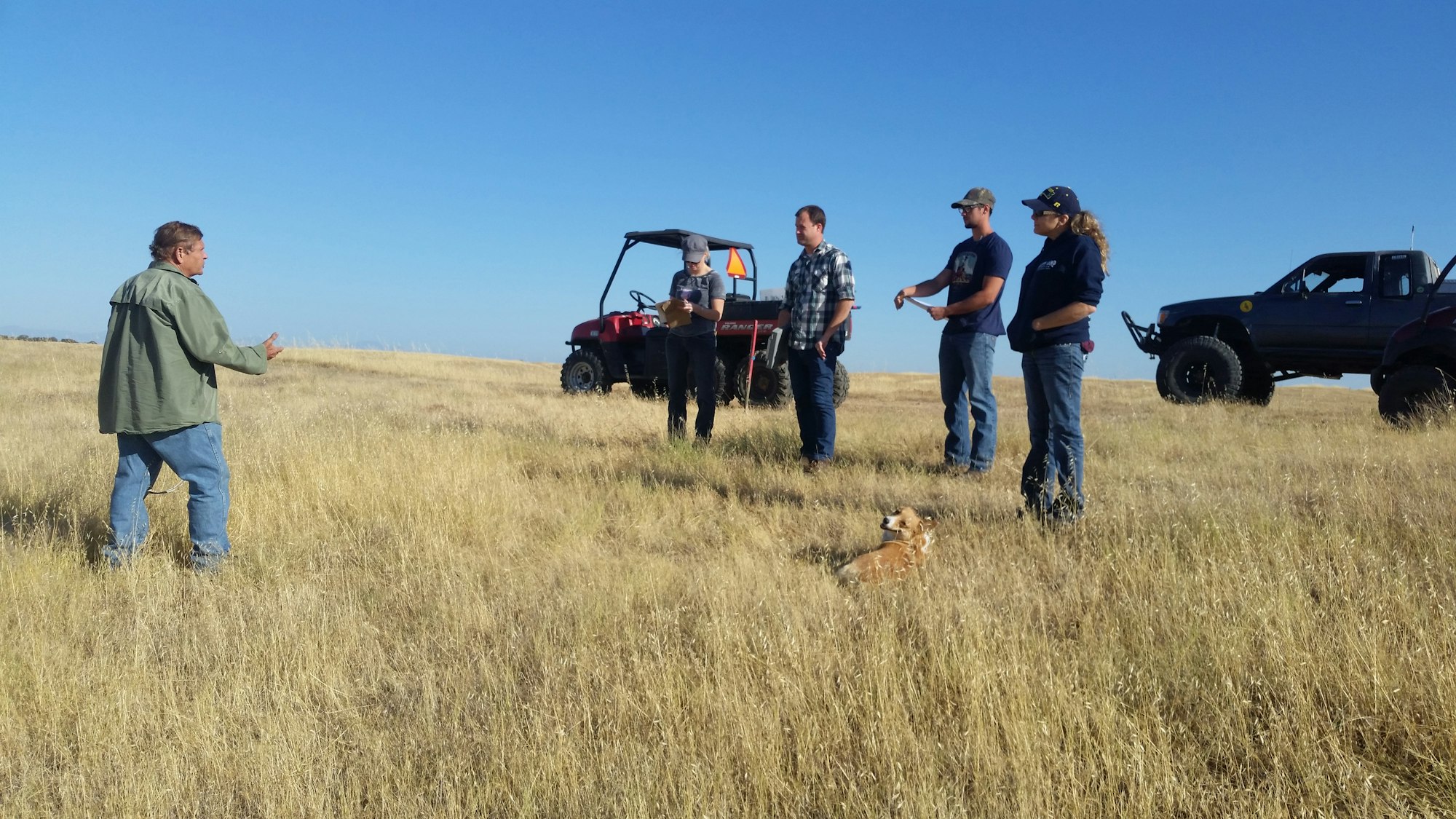 RCDTC are seen standing in a dry grassy field learning to conduct RDM monitoring. Two offroad vehicles are seen in the background.