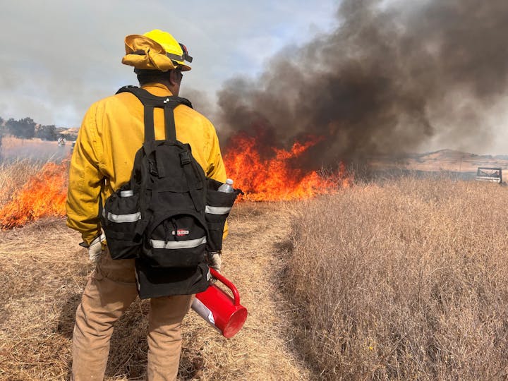 A firefighter stands before a wildland fire, ready to respond with protective gear and equipment.