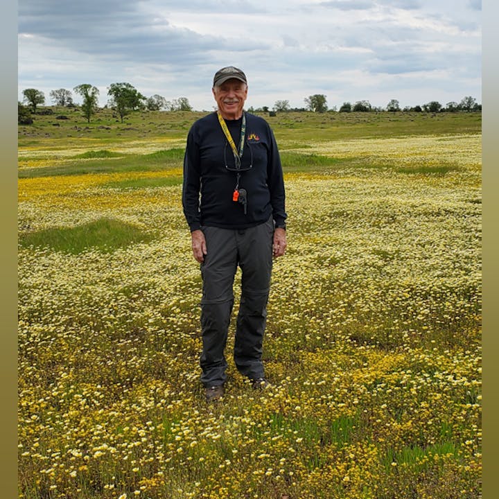 A person stands smiling in a field of wildflowers under a cloudy sky.