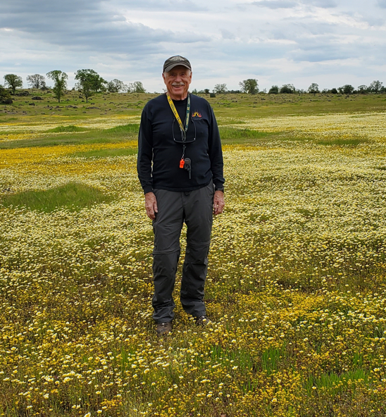A person stands smiling in a field of wildflowers under a cloudy sky.