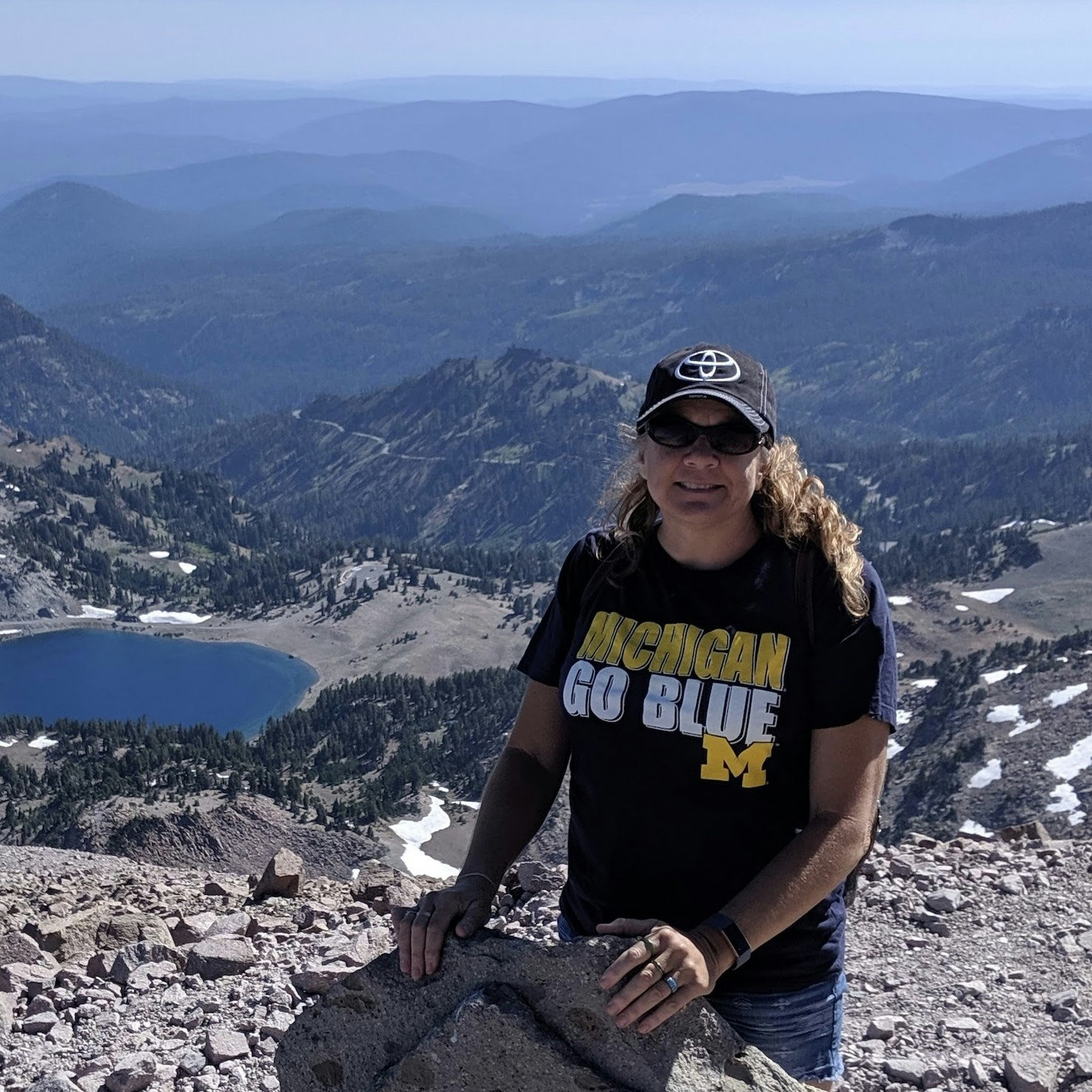 May contain: nature, outdoors, wilderness, clothing, t-shirt, face, head, person, photography, portrait, rock, mountain, mountain range, peak, adventure, hiking, leisure activities, scenery, accessories, sunglasses, lake, water, cap, hat, baseball cap, and pants