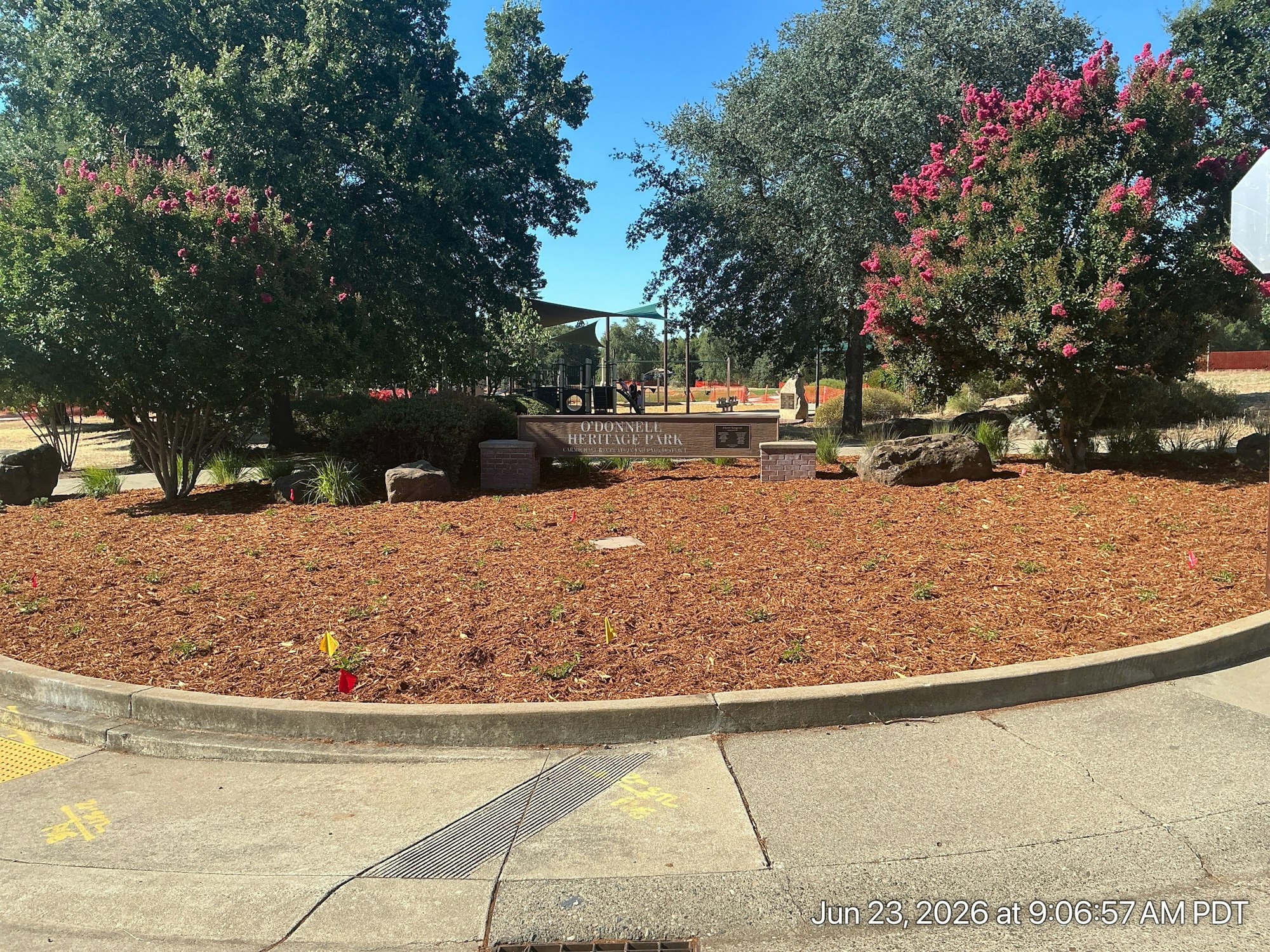 A park landscape featuring trees, flowerbeds with colorful flags, and a sign for O'Donnell Heritage Park.