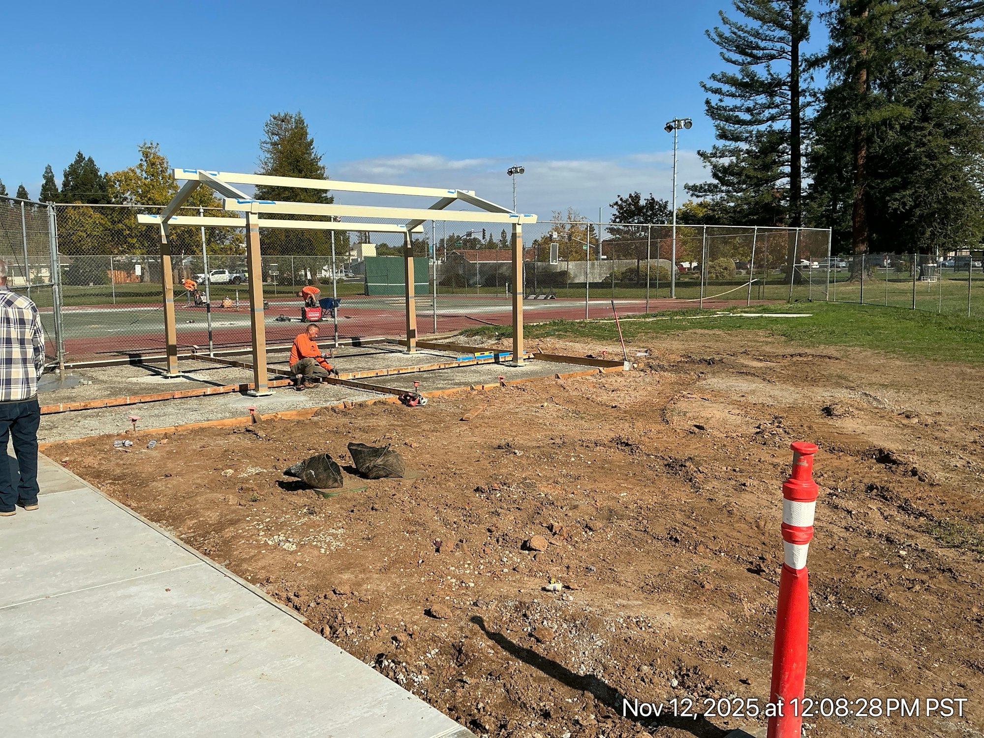 A construction site with workers building a structure next to a fenced area, likely a sports facility, on a clear day.