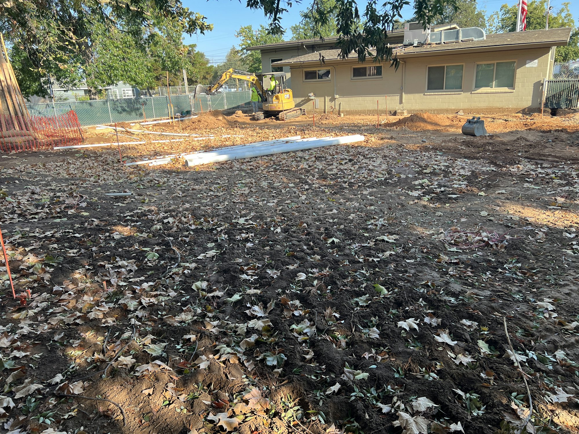 Construction site with dirt ground, fallen leaves, pipes, and an excavator near a building.