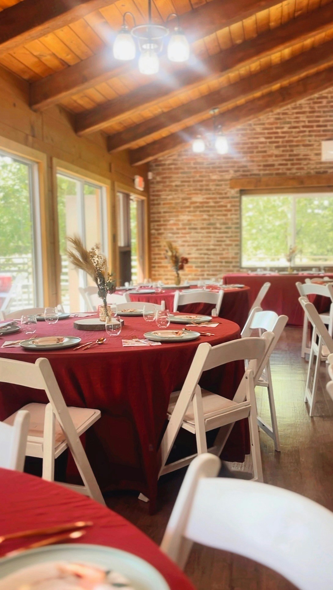 Tables set for an event with red tablecloths, white chairs, and floral centerpieces in a rustic room.