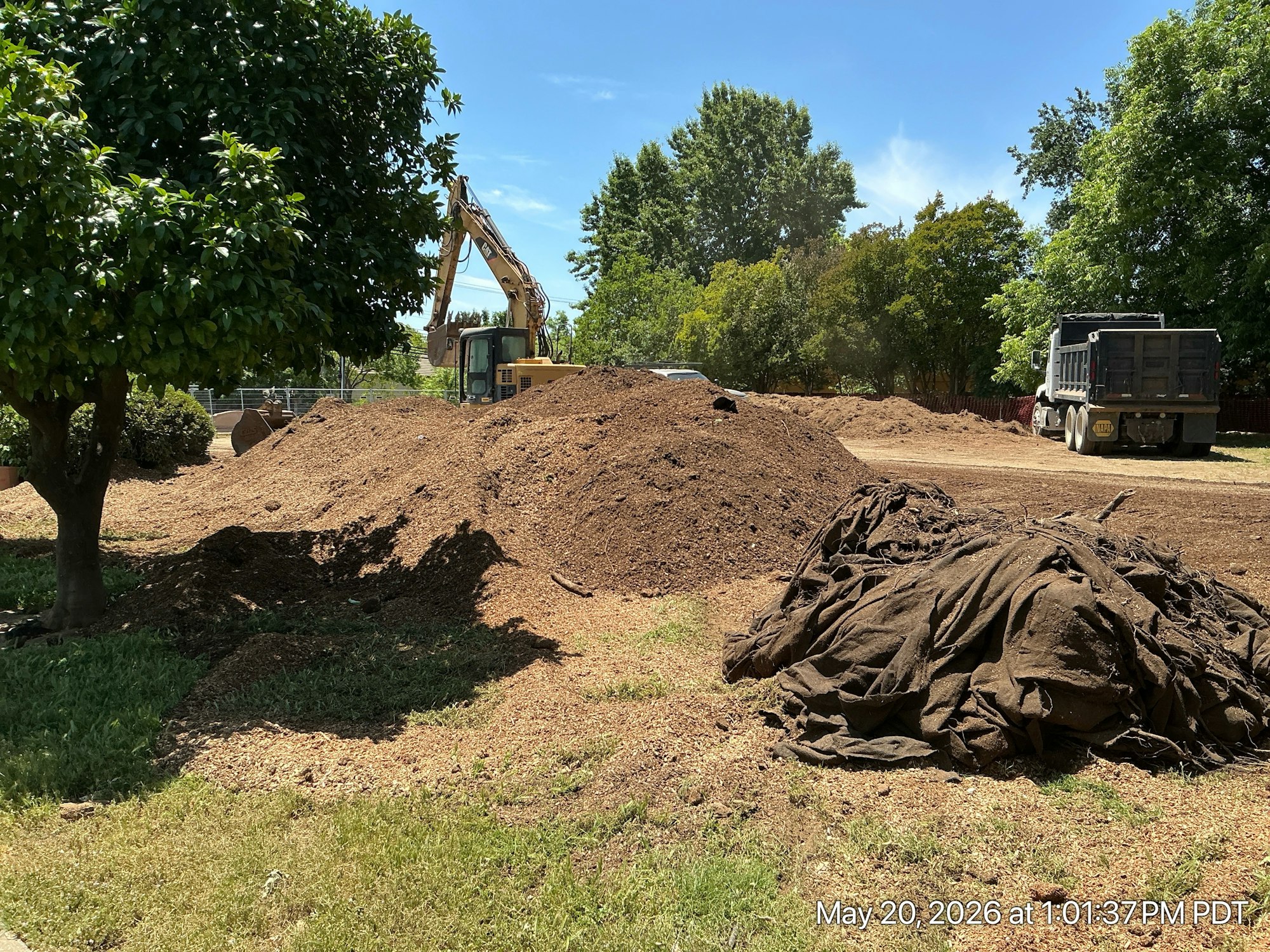 The image features a construction site with large piles of mulch, a tree, and heavy machinery in the background under a clear sky.