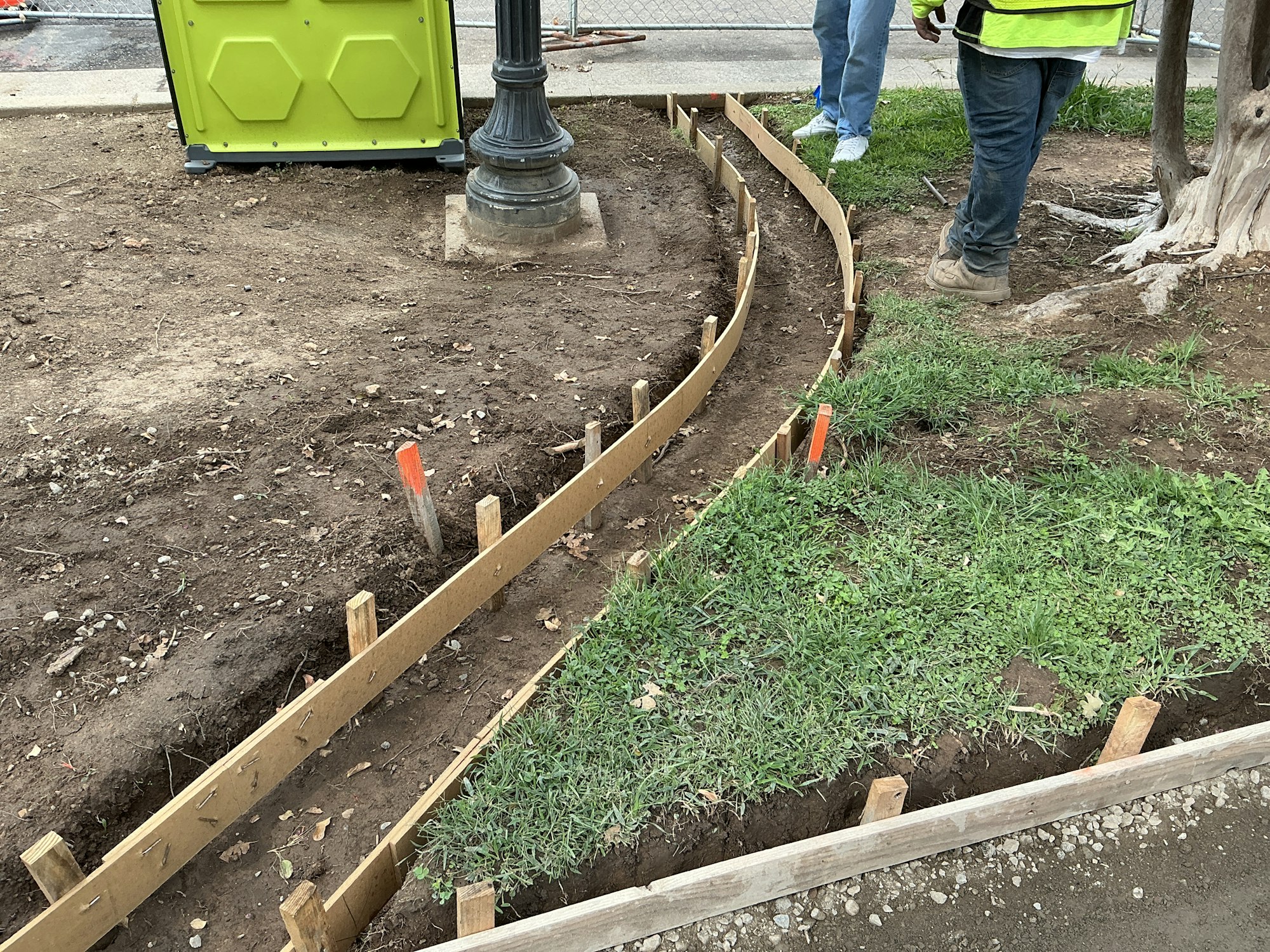 Construction site with wooden formwork for a concrete path, a lamppost, and a green portable toilet. Workers visible.