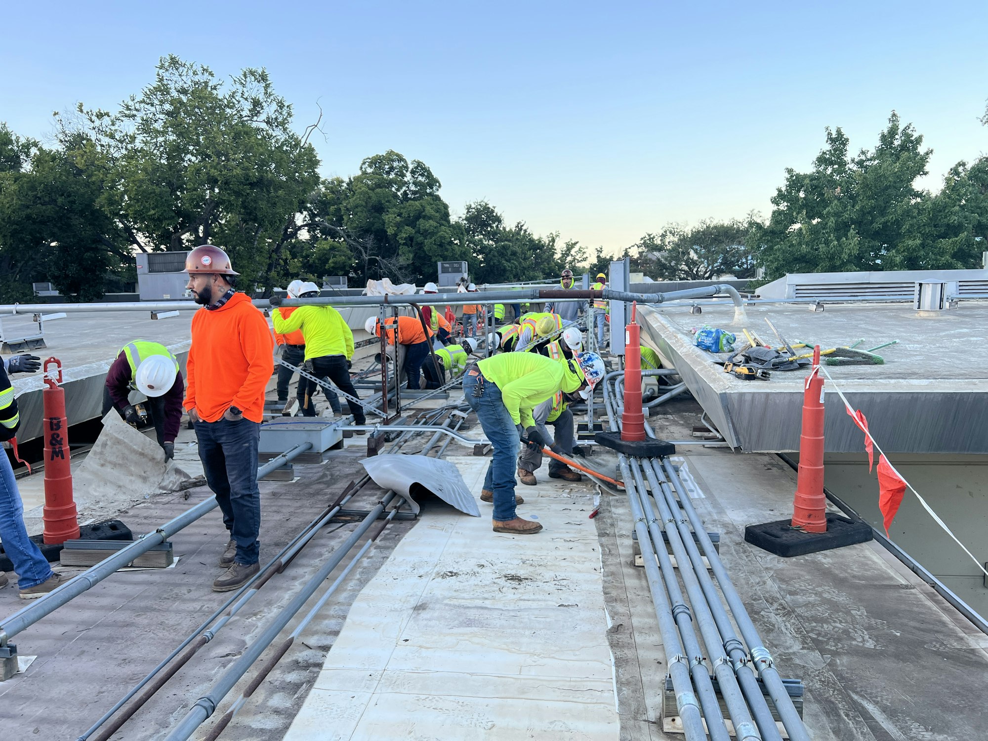 Construction workers in bright safety gear are working on a rooftop, with tools and equipment around.