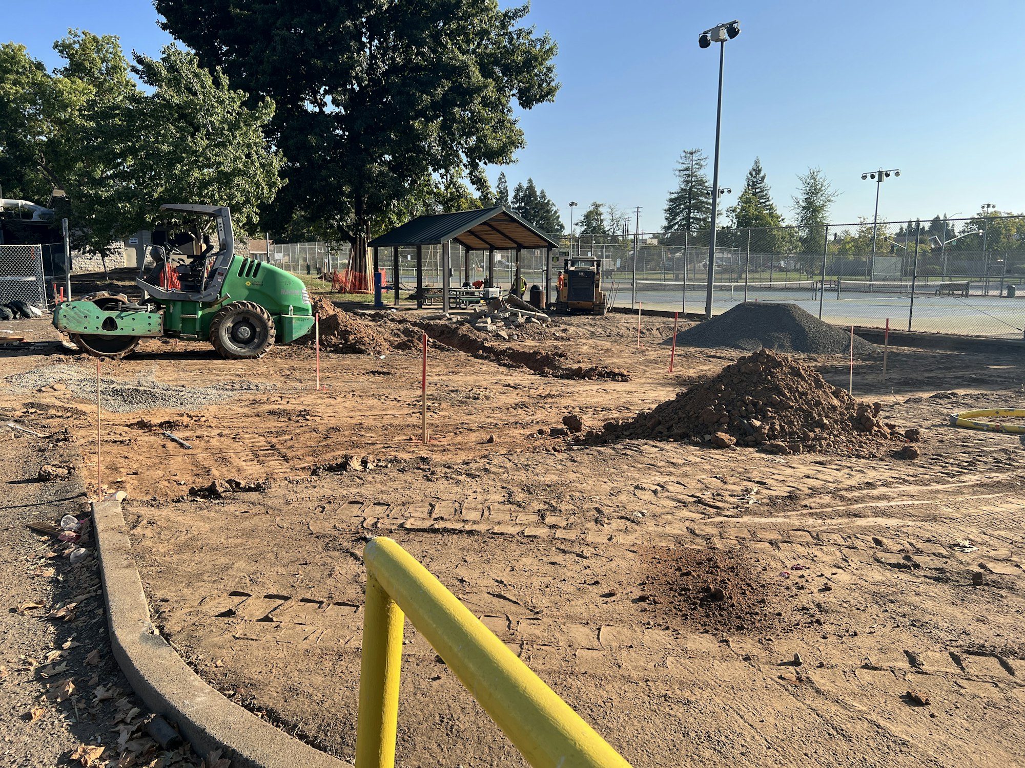 A construction site with a green roller, piles of dirt, and fencing near a tennis court on a sunny day.
