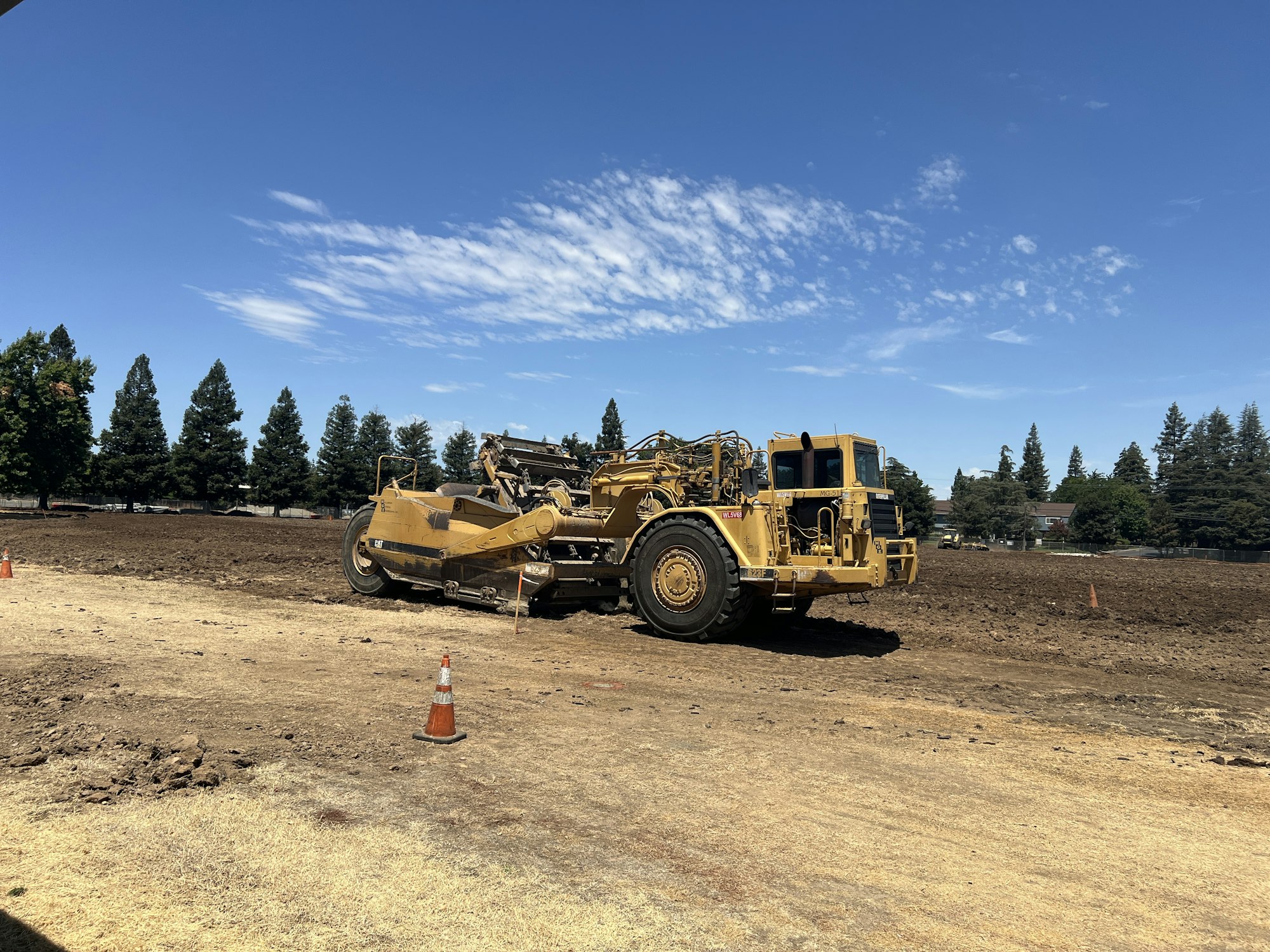 A yellow construction vehicle on a dirt site with scattered traffic cones and trees in the background.