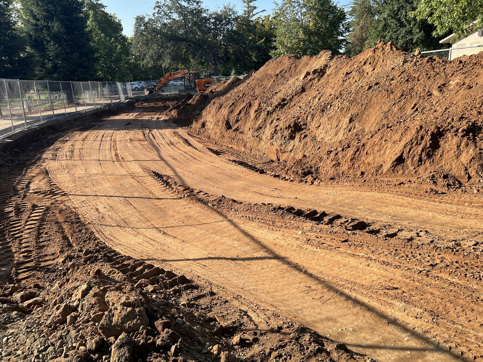 A construction site with a dirt path, a digger, and mounds of earth beside a fence, surrounded by trees.