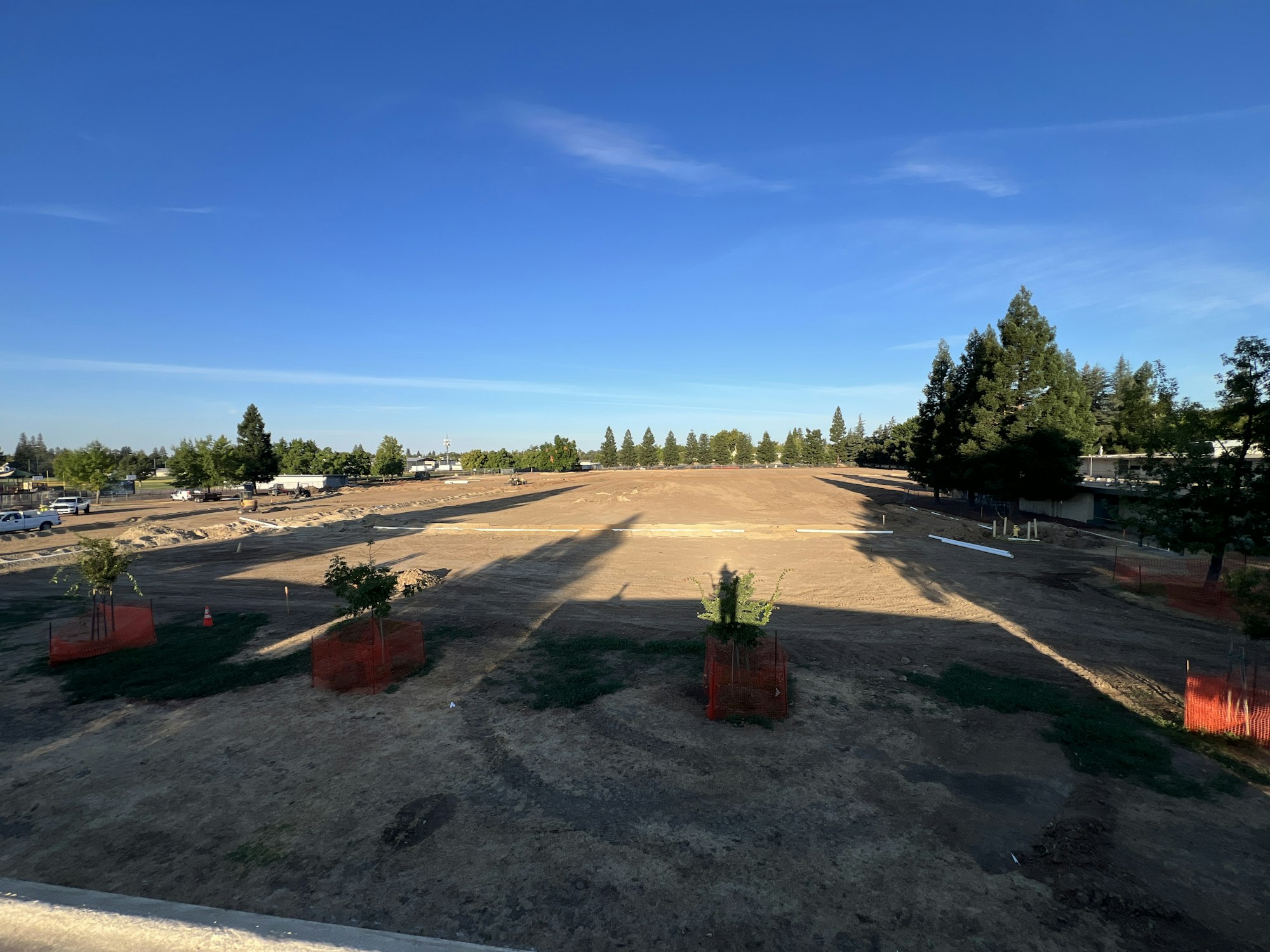 Open field with construction materials, small fenced trees, and distant vehicles under a clear blue sky.